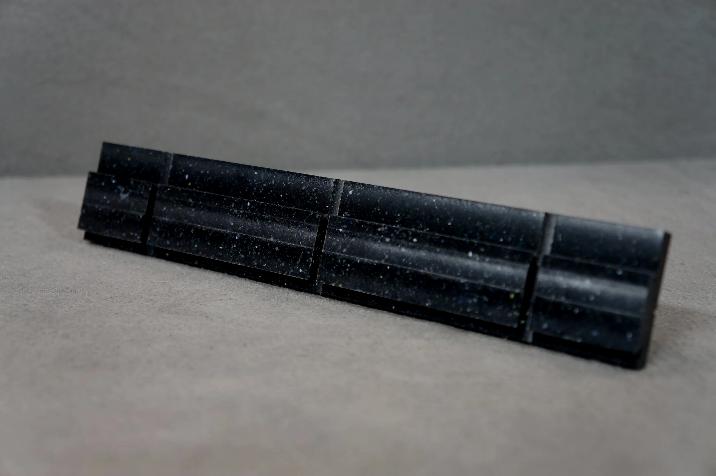 A black rectangular object with speckles and a textured surface, placed on a flat, light gray surface with a gray background.