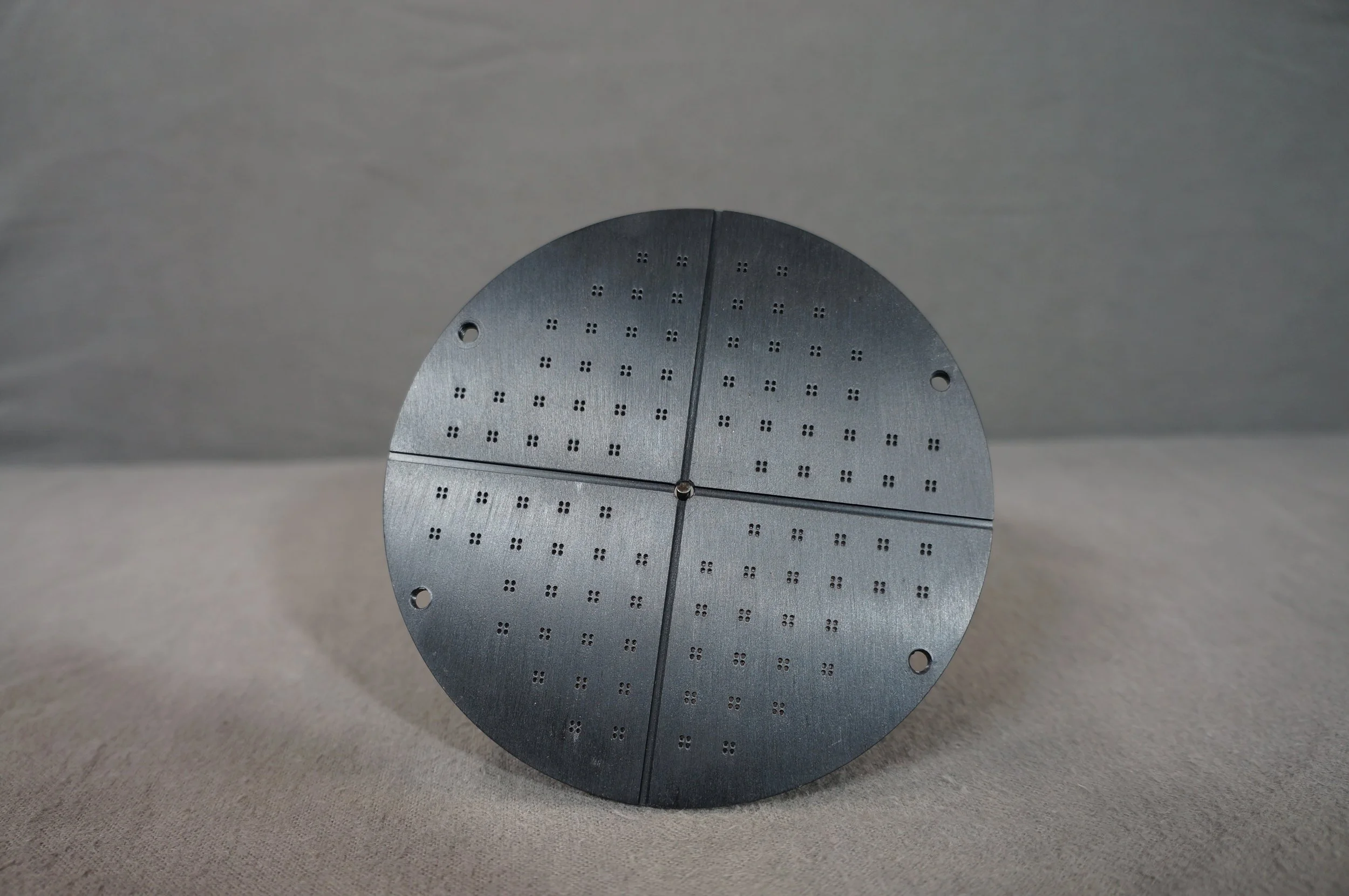 A metallic circular object with multiple small holes arranged in a grid pattern, divided into four quadrants by two intersecting lines, lying on a gray surface.