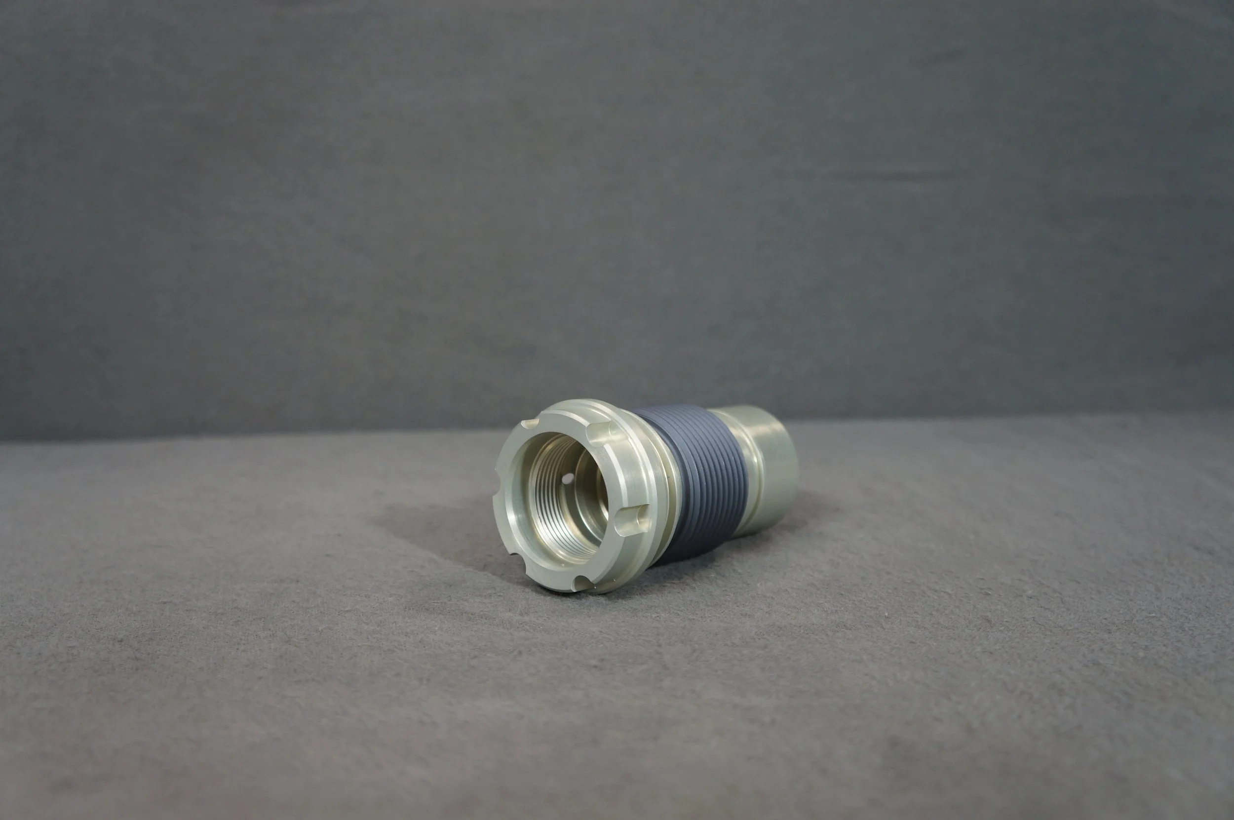 A metallic cylindrical object with a ridged rubber ring around its middle, lying on a gray surface against a dark gray background.