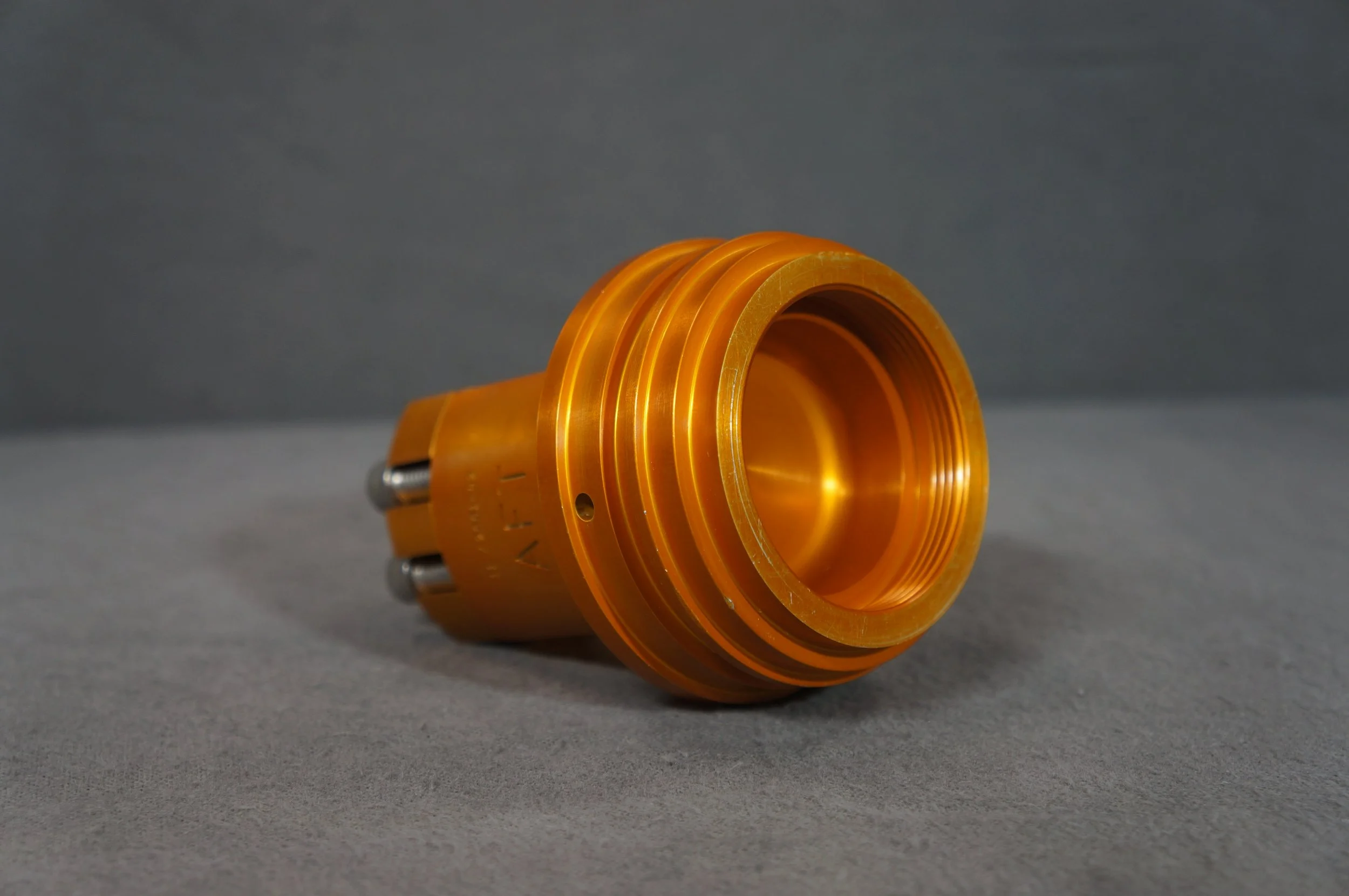 Orange metallic mechanical part with grooves and threaded interior resting on a gray surface with a gray background.