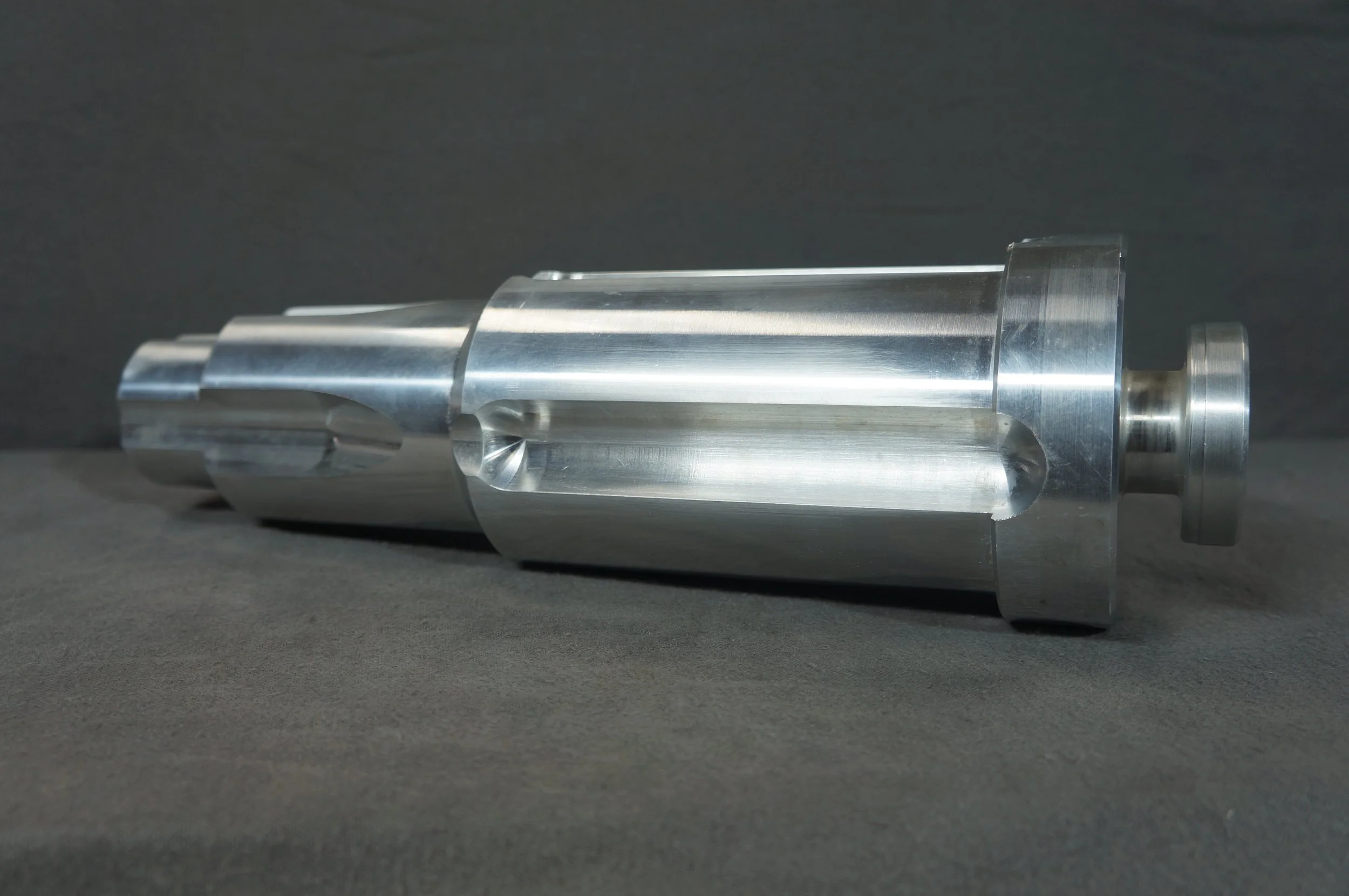 A shiny, metallic, cylindrical machine part with a flat end and grooves, lying on a dark surface against a dark background.