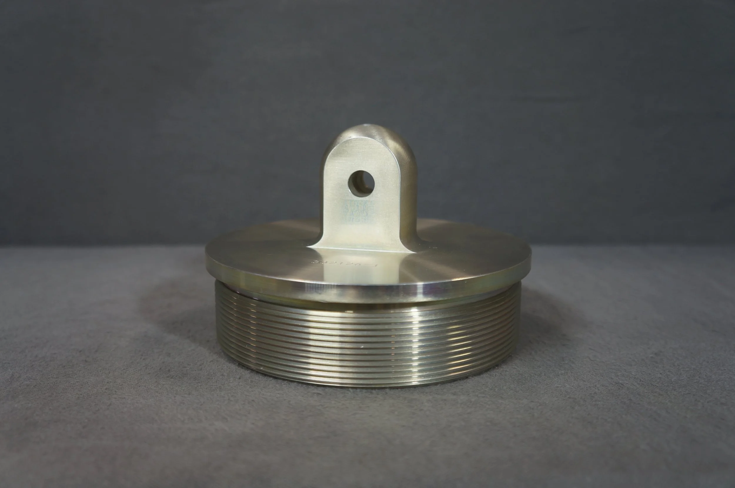 A close-up view of a metallic flange with a threaded base and a central hole, positioned on a flat surface against a gray background.