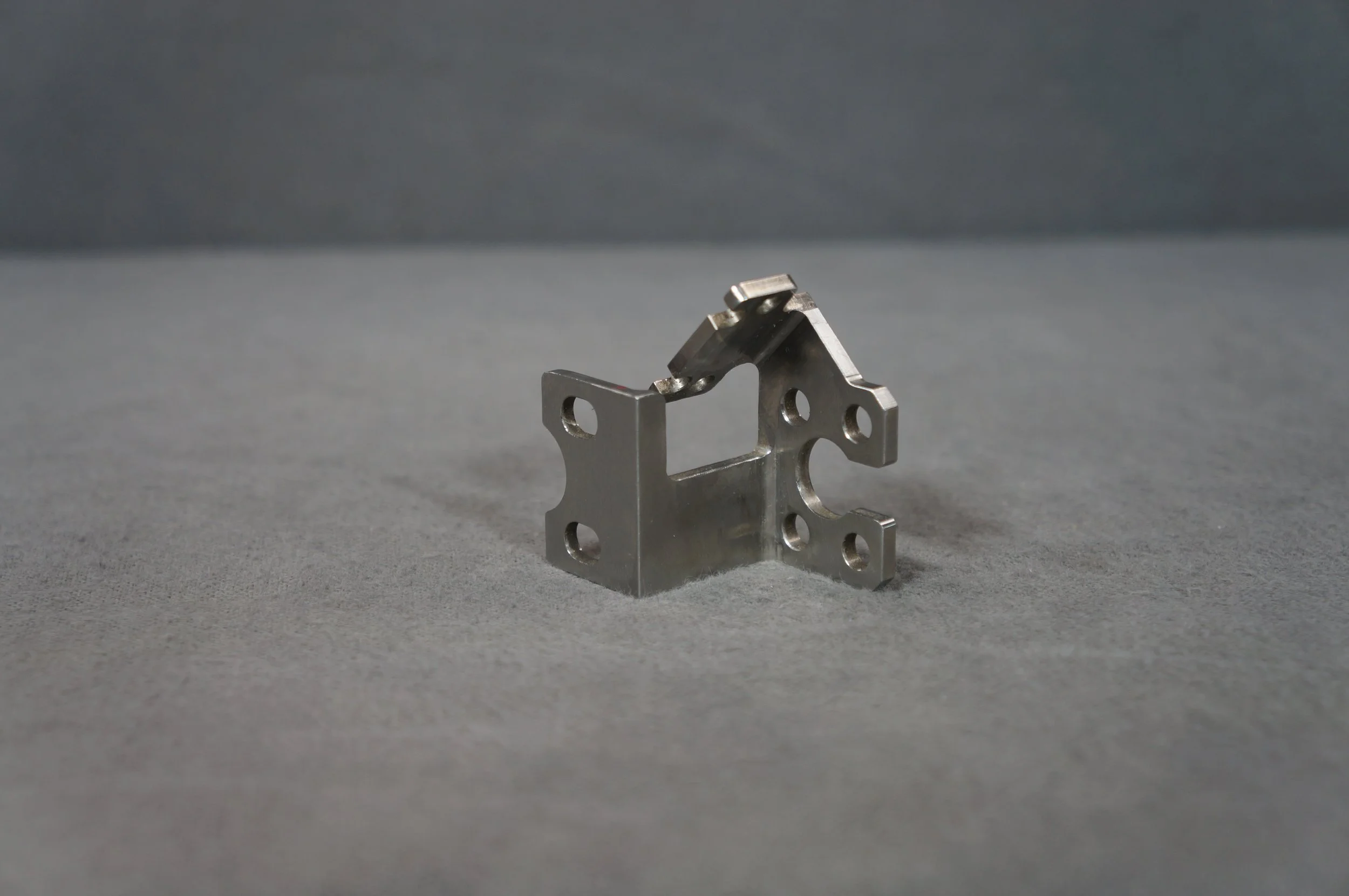 A small metallic mechanical part with multiple holes and a bent shape, positioned on a gray surface against a dark gray background.