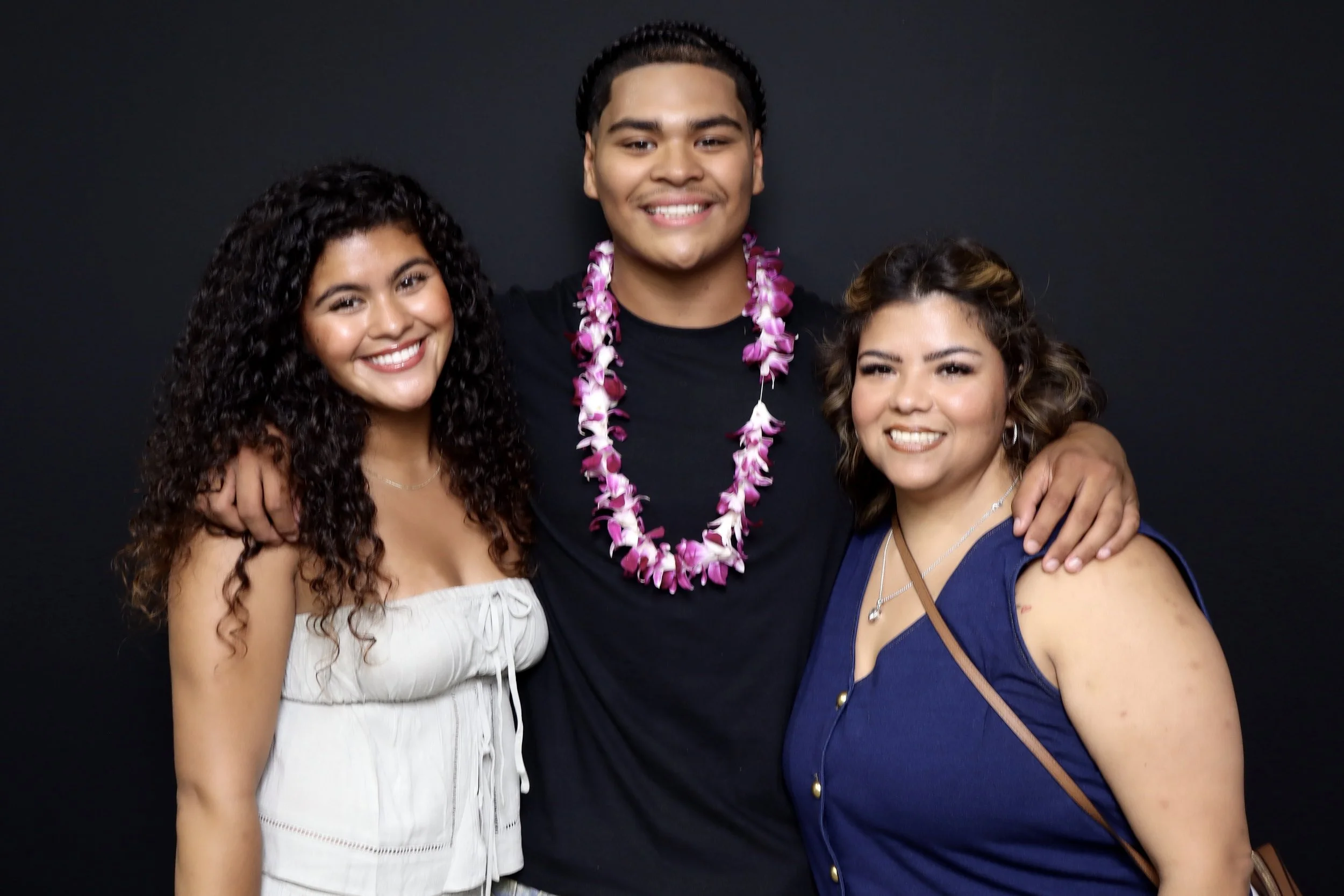 Three young adults posing closely together against a dark background, smiling for the camera. The person in the middle is wearing a pink flower lei.