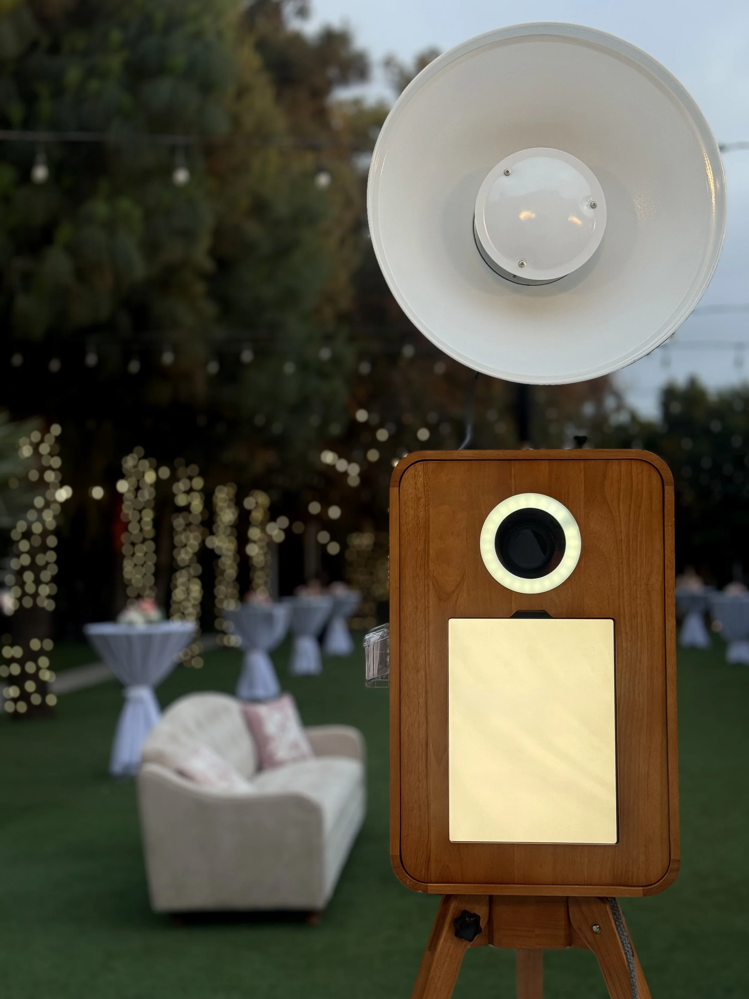 A wooden photo booth with a light ring and a blank sign in an outdoor setting with string lights and garden tables in the background.