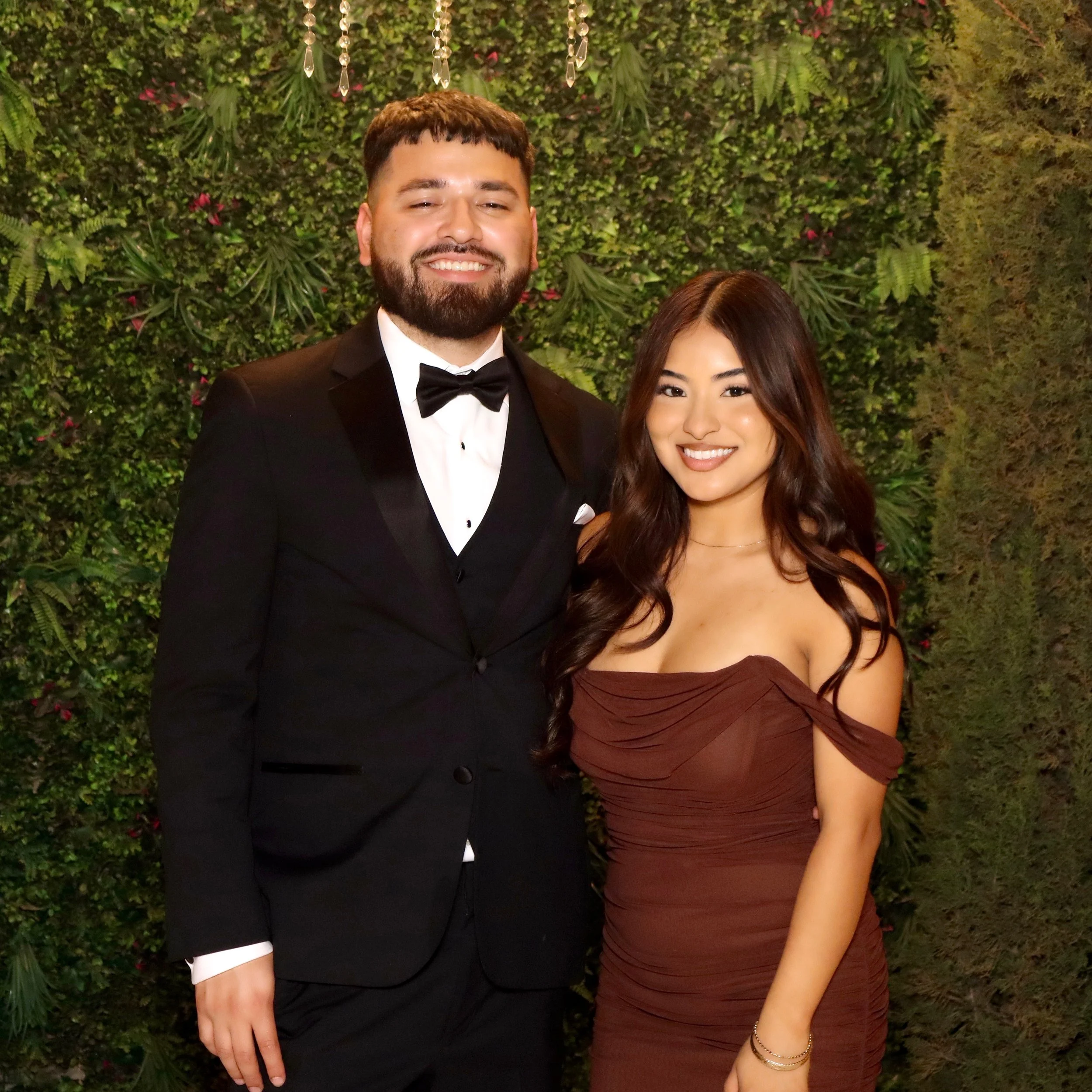 A smiling man in a tuxedo standing next to a smiling woman in a strapless dress, both posing in front of a green, leafy wall with hanging crystals.