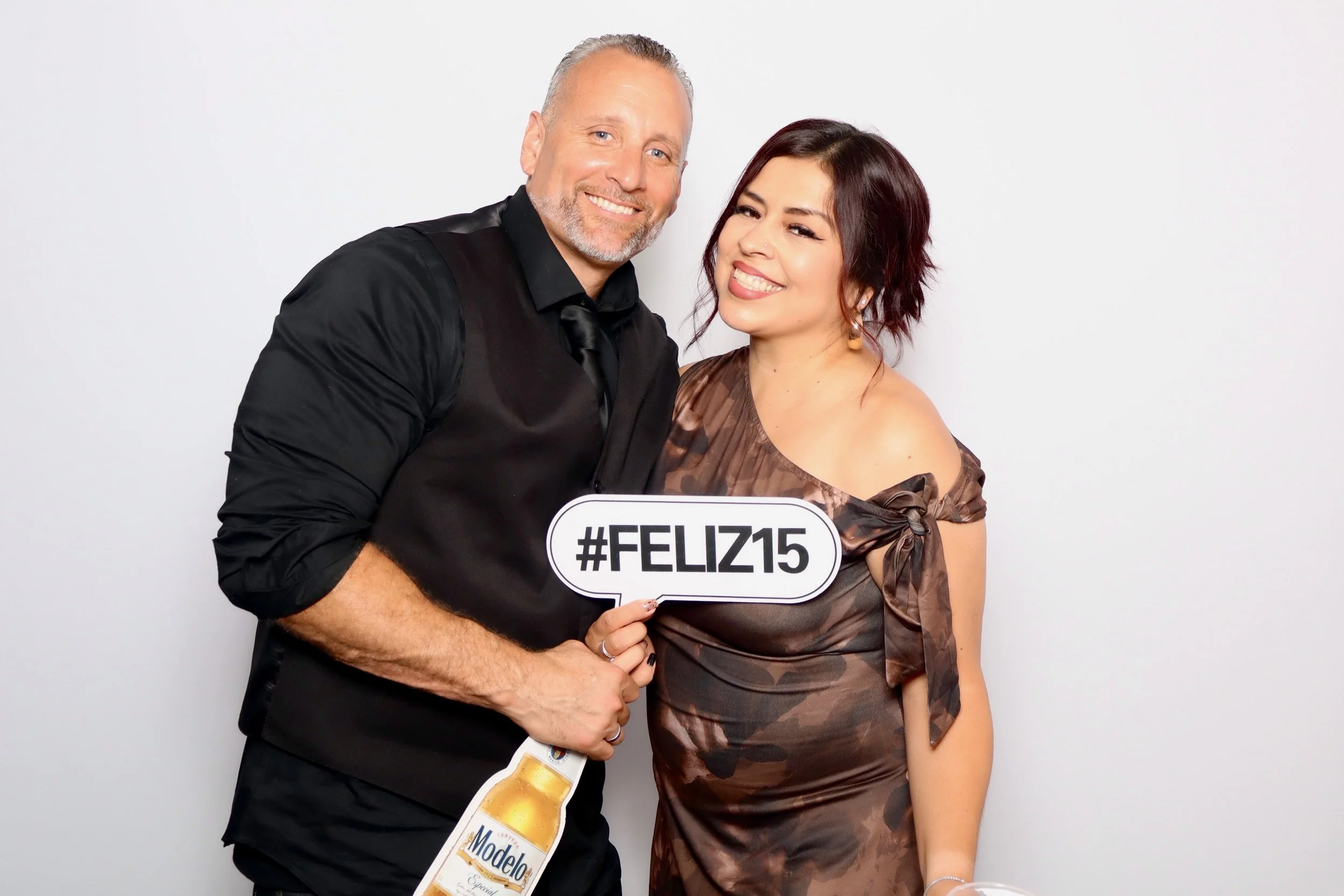 A happy man with gray hair and beard and a woman with short dark red hair and a dress holding a sign that says #FELIZ15, a beer bottle cutout, and a glass of beer, posing against a plain white background.