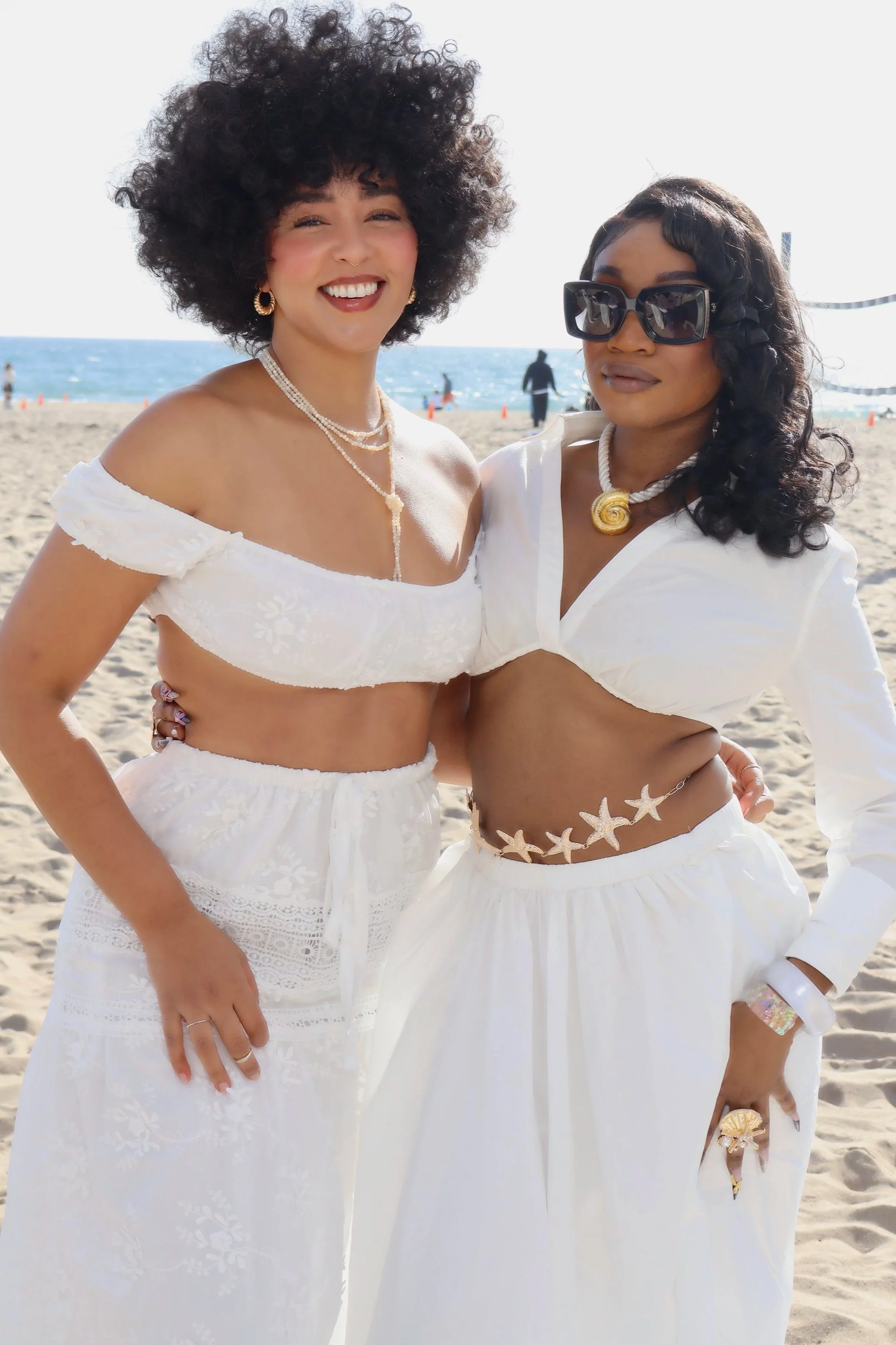 Two women dressed in white beachwear, standing on a sandy beach with the ocean and a few people in the background. The woman on the left has curly hair, is smiling, wearing earrings, layered necklaces, and a white off-shoulder top. The woman on the r