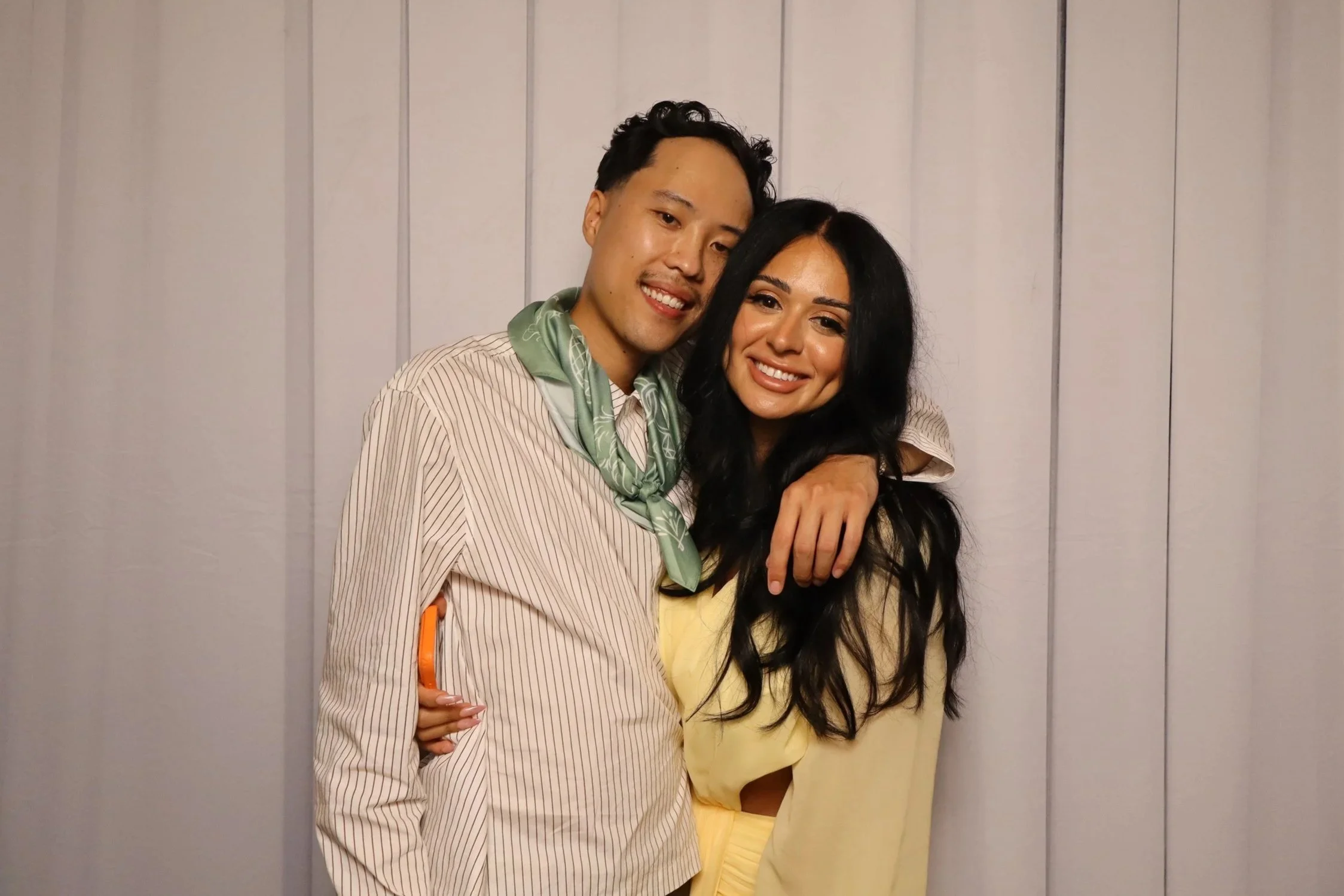 A smiling young man and woman embrace each other, standing in front of a plain, light-colored wooden wall. The man has dark, curly hair and is wearing a striped shirt with a green neck scarf, while the woman has long, dark hair and is dressed in a li