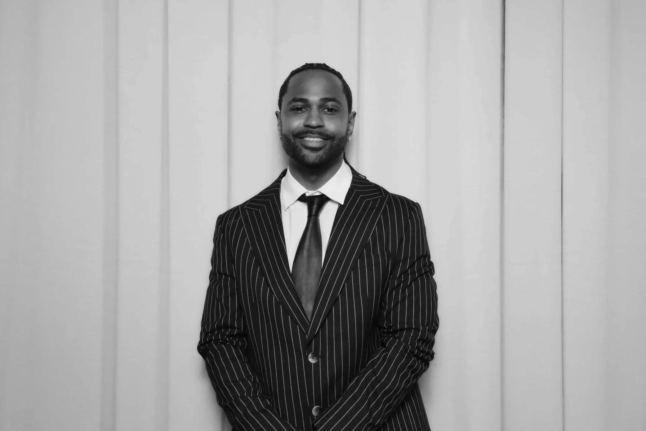 A man in a pinstripe suit and tie standing against a light wall with vertical lines.