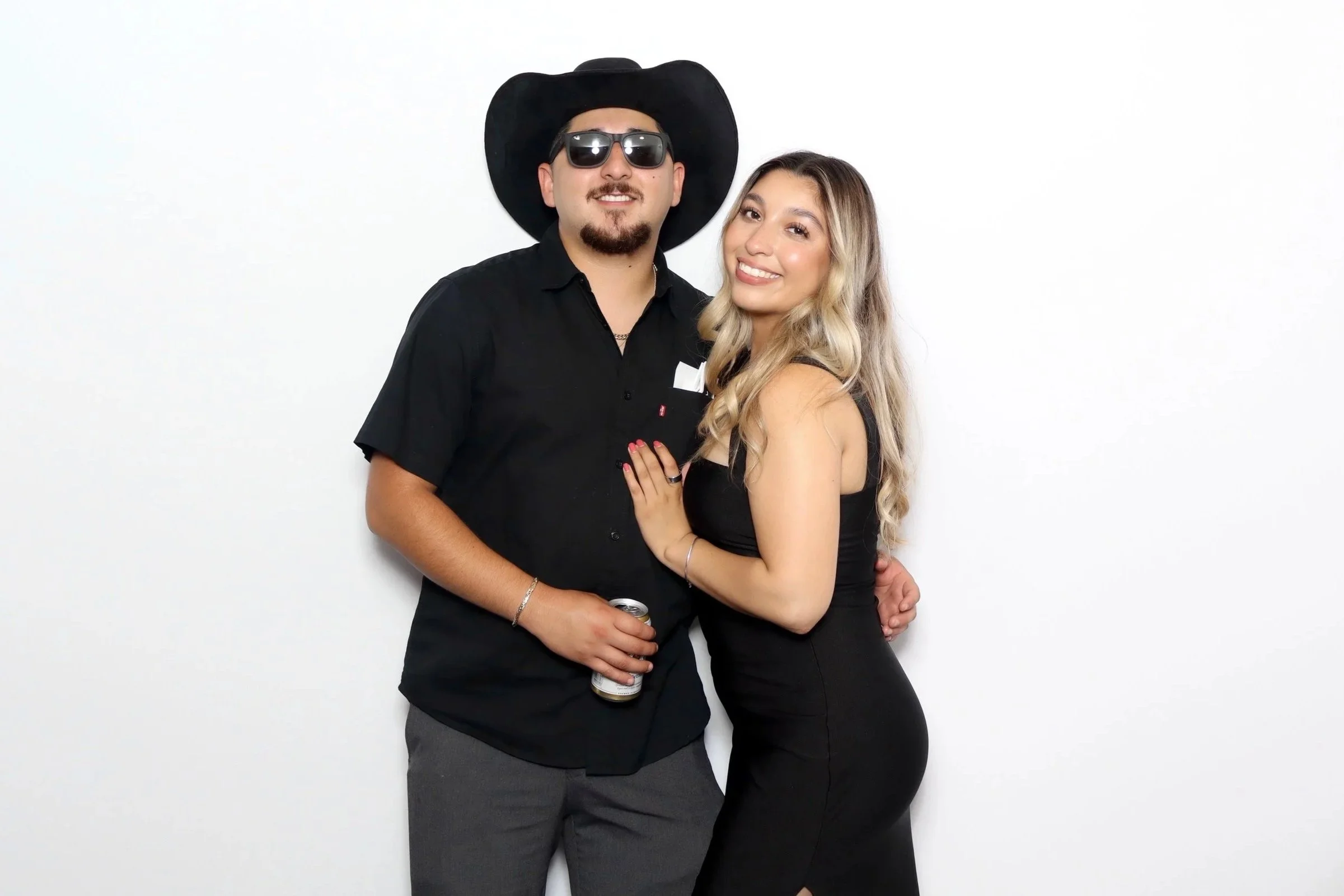 A man wearing sunglasses, a black cowboy hat, and a black shirt stands beside a woman with long wavy blonde hair wearing a black dress. They are smiling and close together, with the woman’s hand on the man's chest. The man is holding a can in his lef