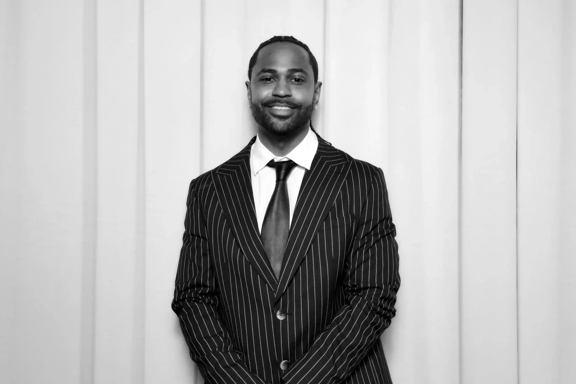 Rapper Big Sean wearing a suit and tie posing smiling against a elegant white background.