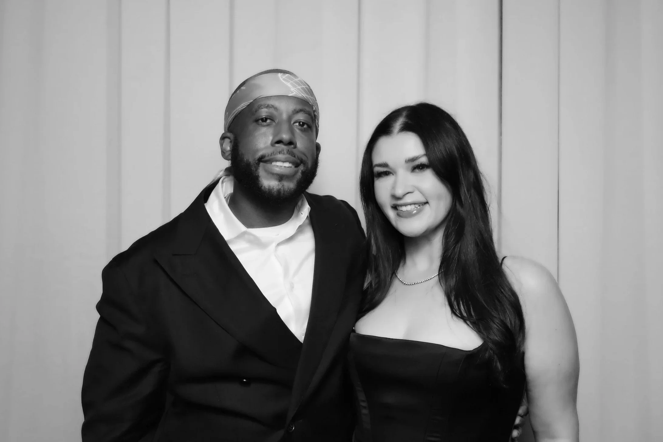 Couple posing dressed formal, the woman has long dark hair and the man is wearing headwear. They are against a draped background with a classic black and white filter known as the "glam filter" or the "kardashian filter".
