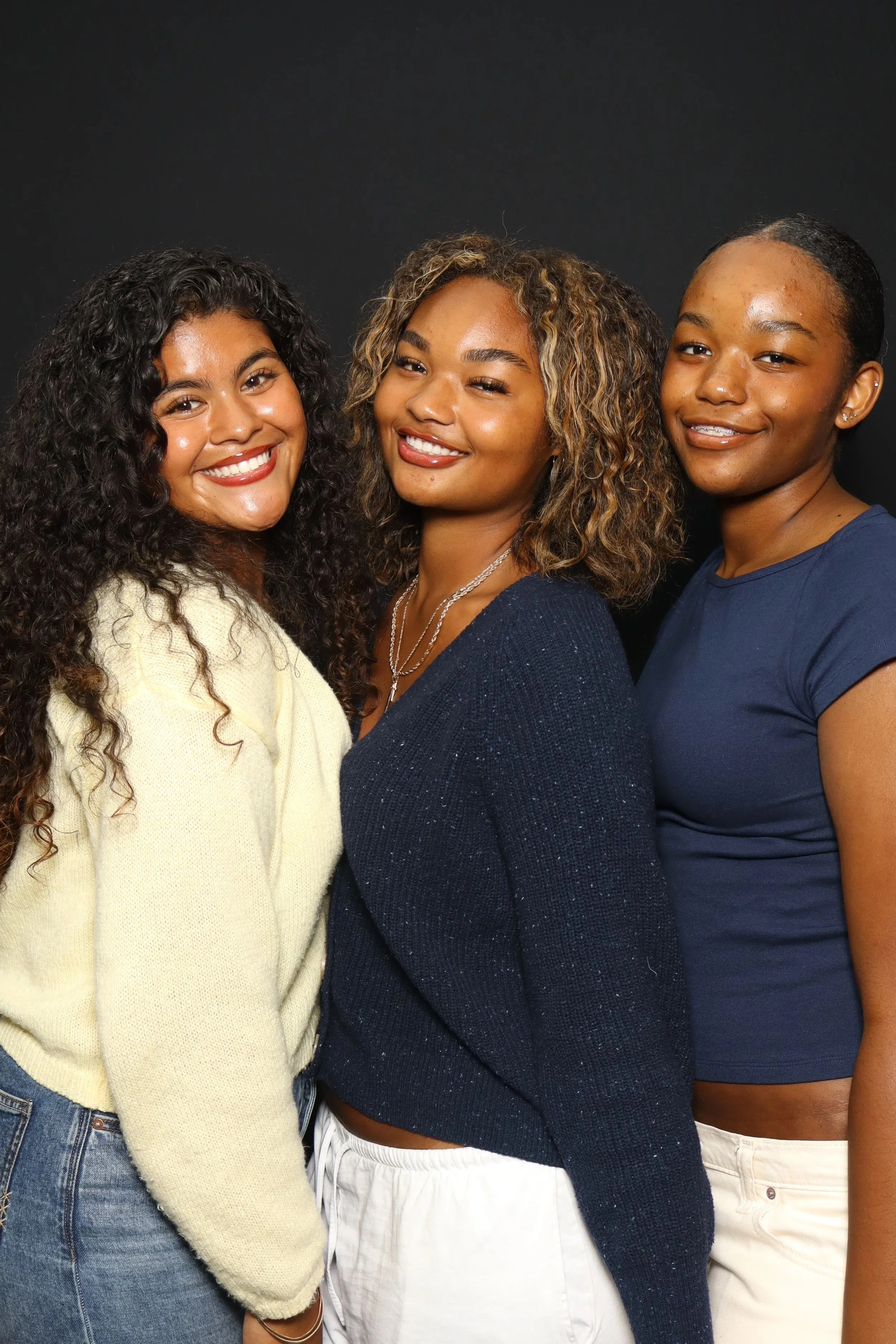 3 girls posing together smiling against a classic black background. 2 of them have curly hair and one has a slick back pony tail. They are dressed casual in neutral colors.