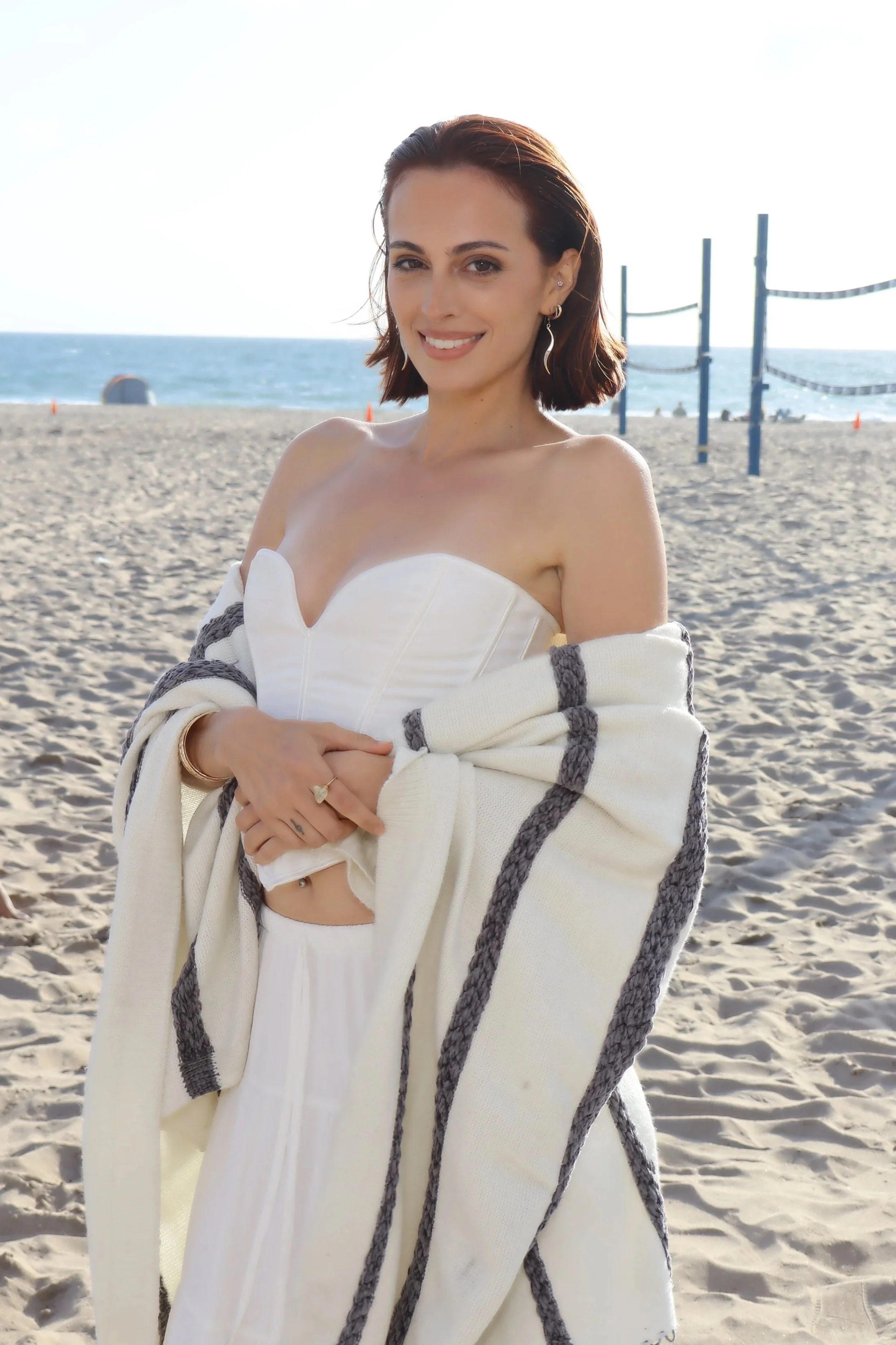 A woman with short hair dressed in white with a cardigan and earrings posed infront of the beach posing and smiling.