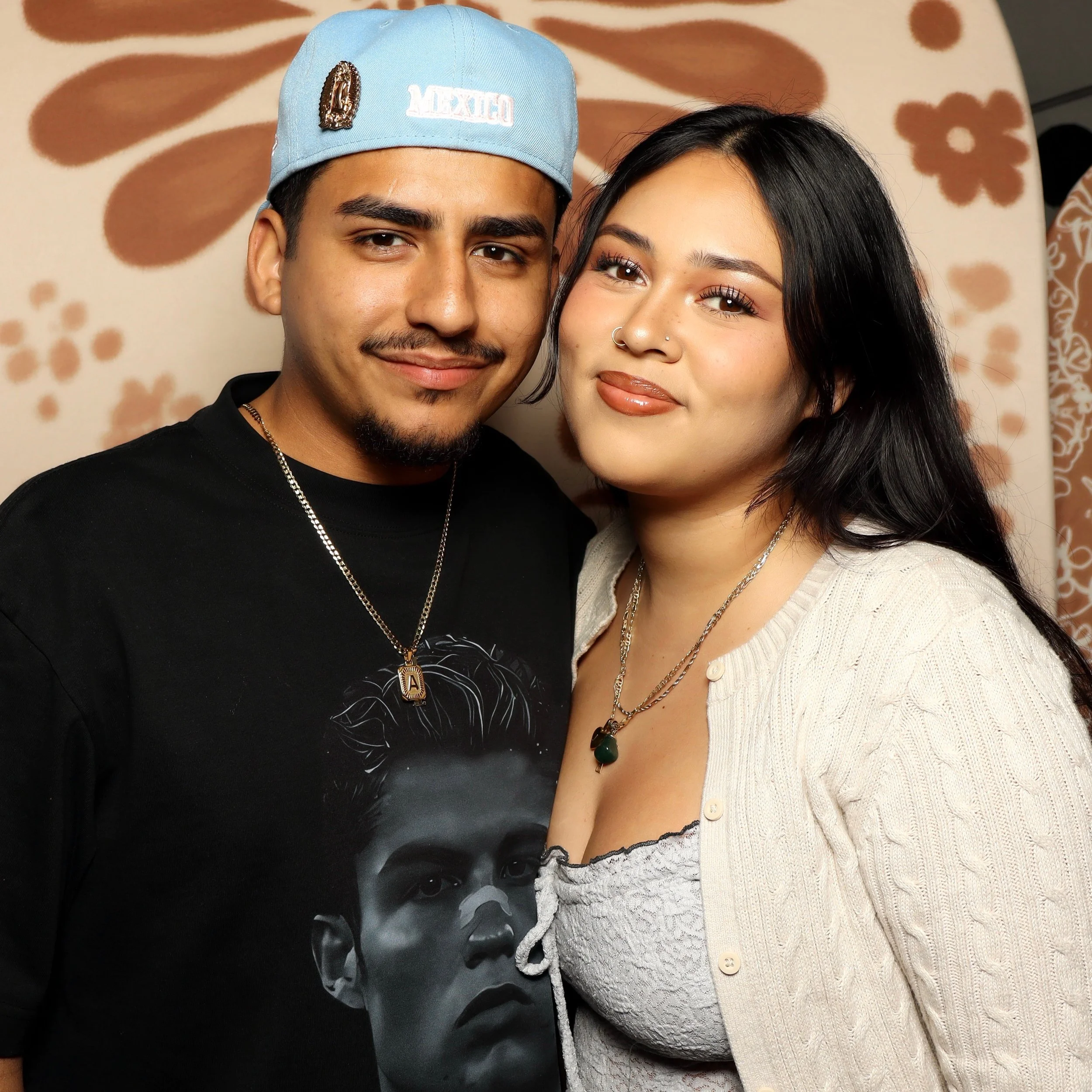 A young man and woman taking a close-up selfie together. The man wears a light blue cap with patches and a black shirt, and the woman has dark hair, makeup, a nose ring, and wears a cream-colored sweater with buttons.