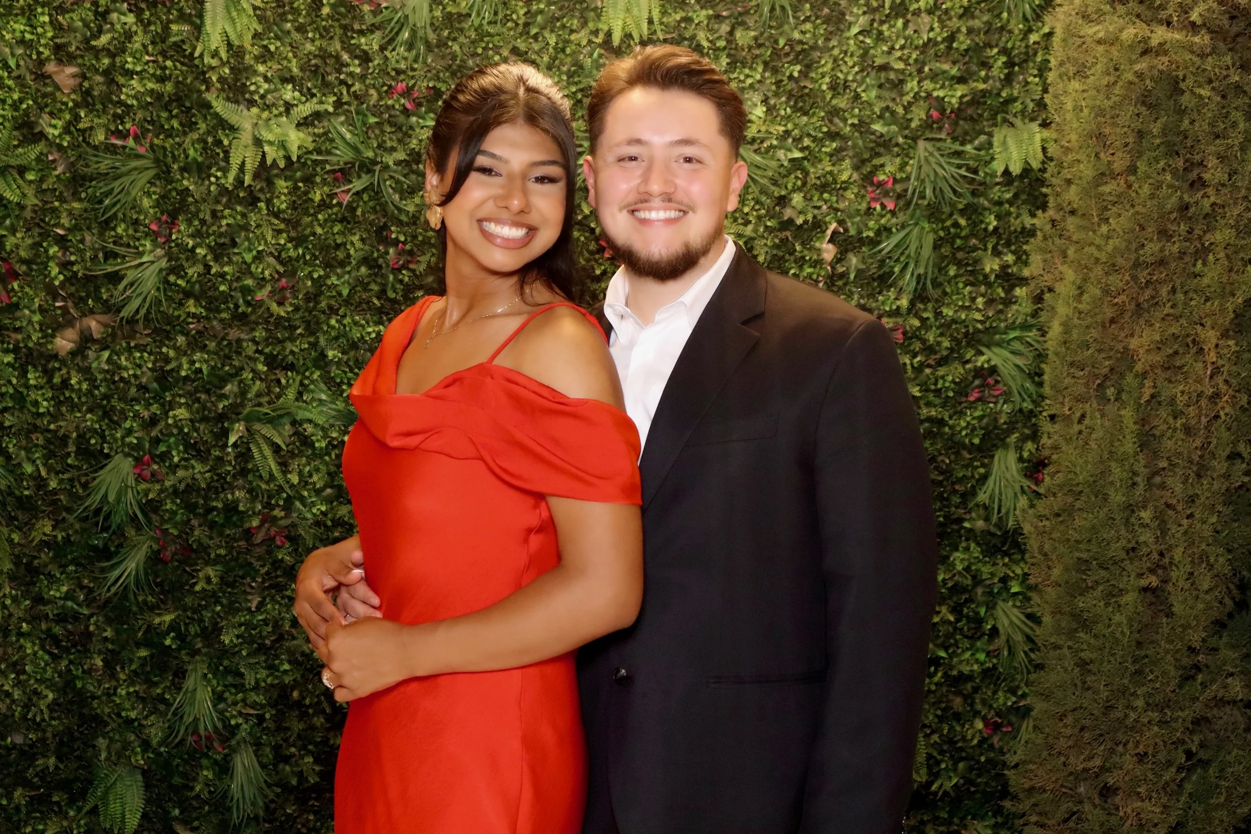 A smiling woman in a red dress and a smiling man in a black suit stand close together in front of a green leafy wall, posing for a photo.