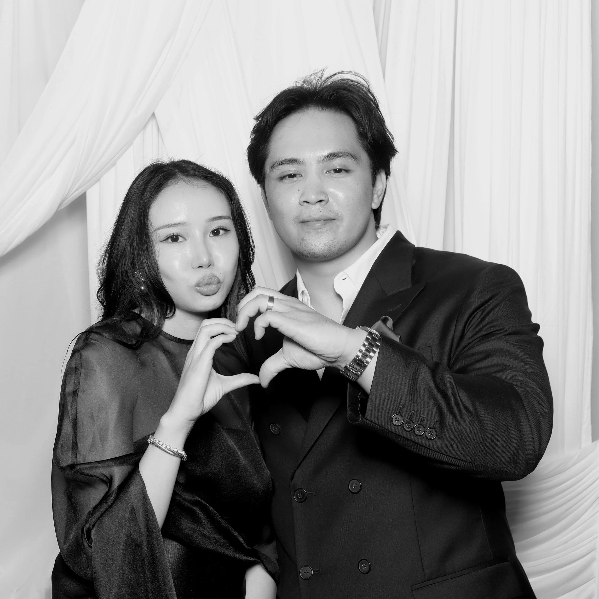 a young woman and man posing against an elegant drape backdrop while making a heart with their hands