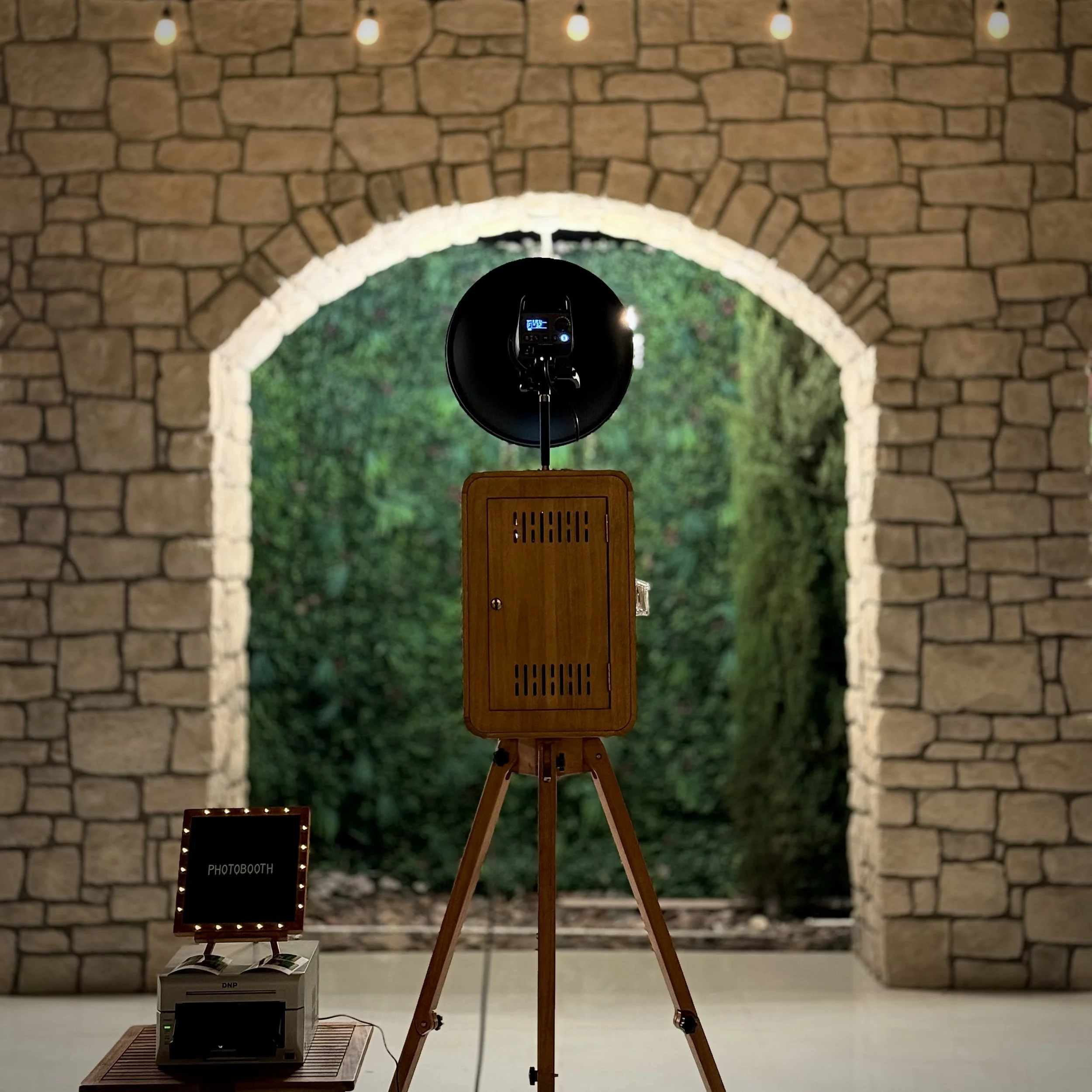 A photo booth setup with a wooden stand, a black camera in a circular holder, a small sign labeled 'PHOTOBOOTH' nearby, and string lights hanging on a brick wall background.