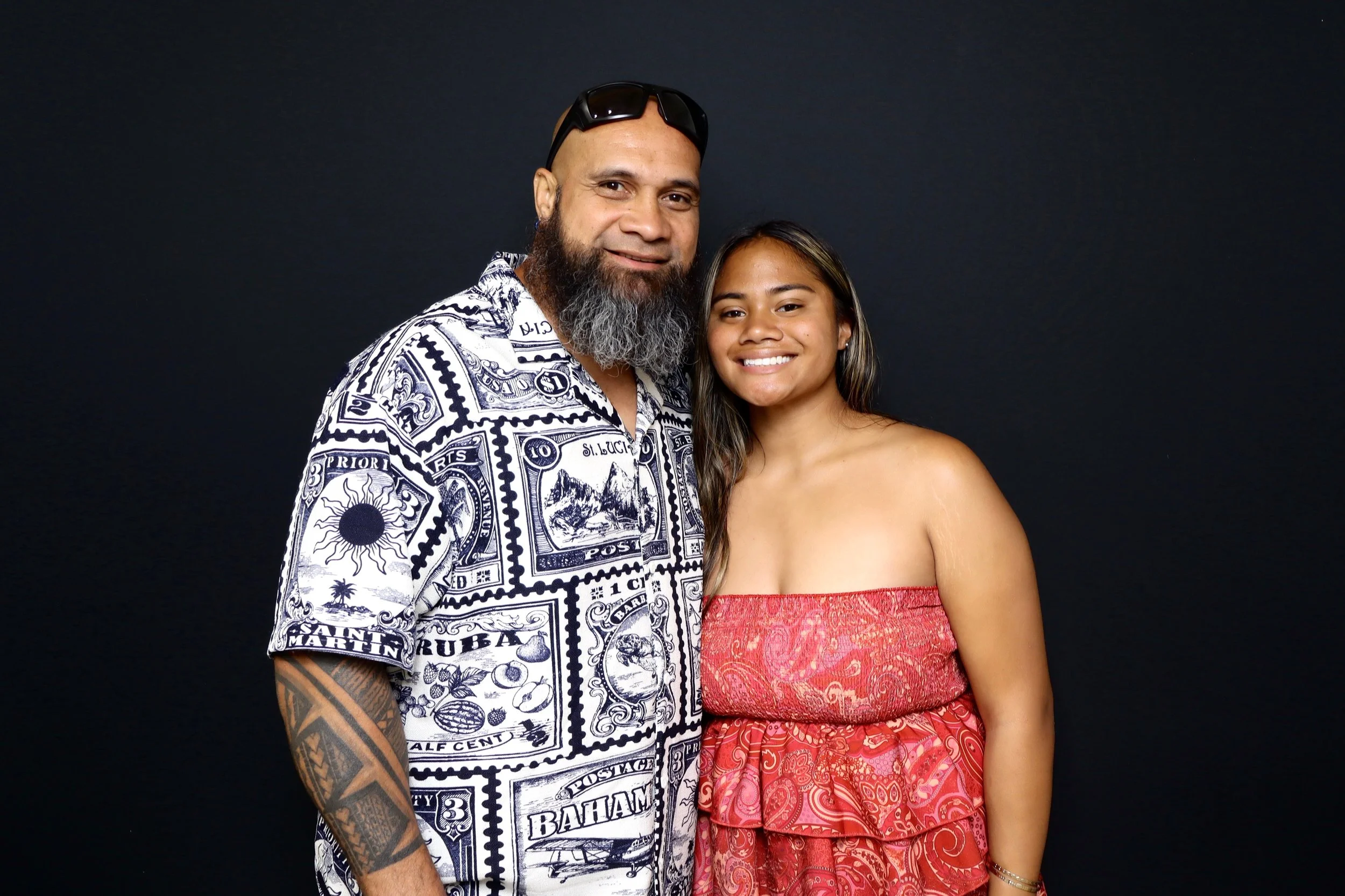 A man with a beard wearing a patterned shirt and sunglasses on his head, standing next to a woman with long hair wearing a strapless red dress, both smiling against a dark background.