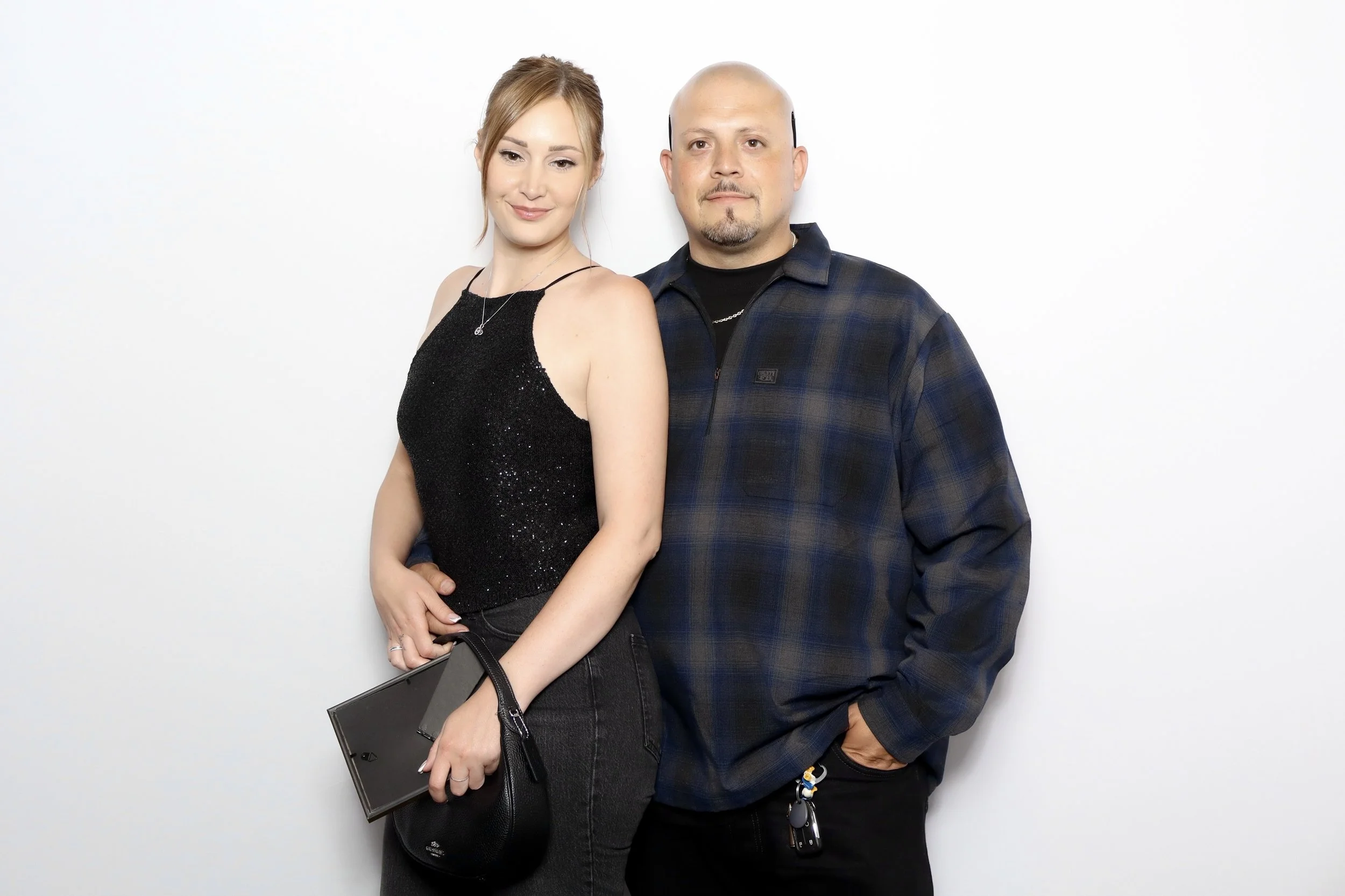 A man and a woman standing close together against a plain white background. The woman has light skin, auburn hair, and is wearing a black sleeveless top with a sparkly texture, holding a black purse. The man has light skin, a bald head, and is dresse
