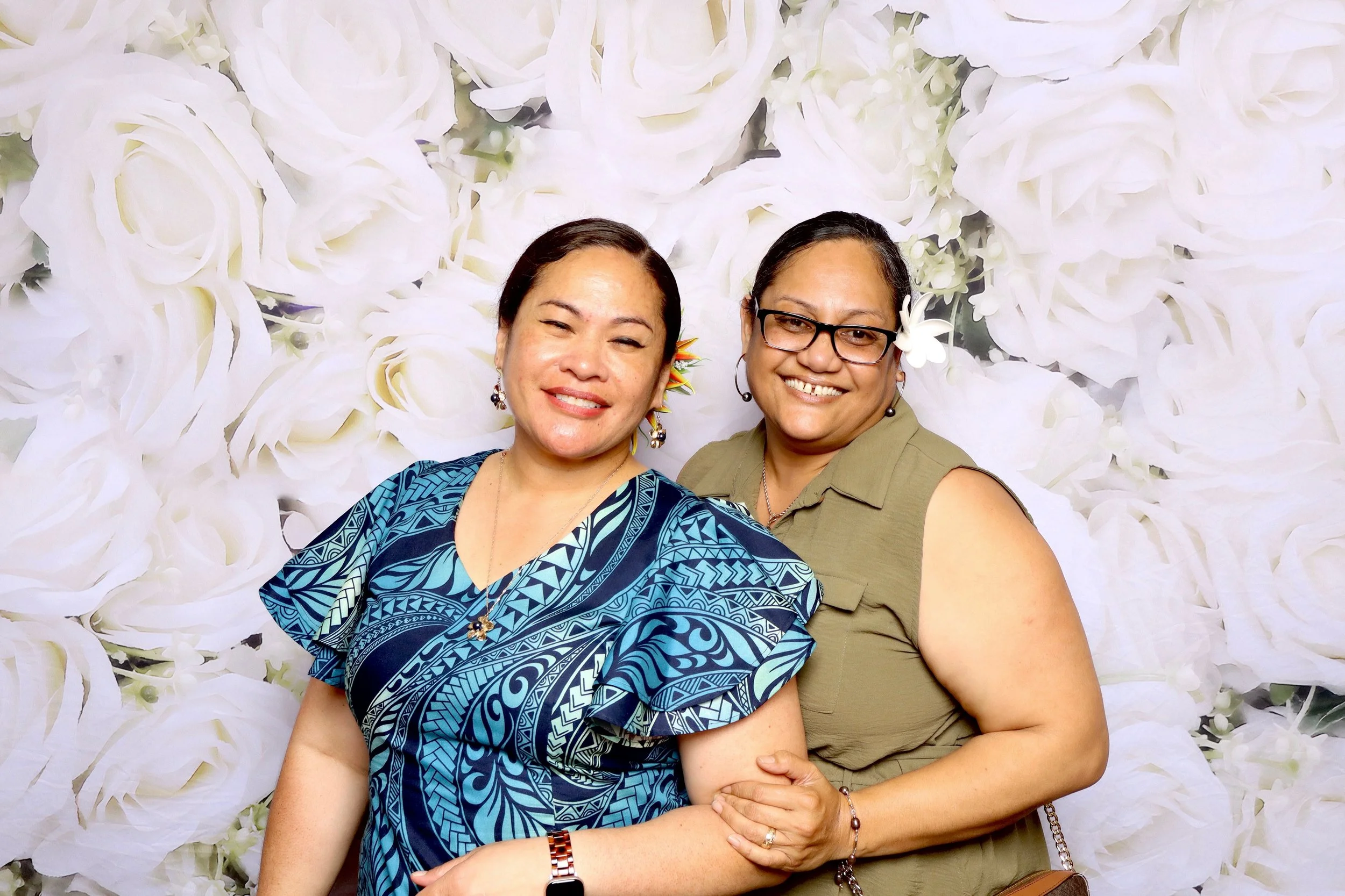 Two women smiling, one with glasses and a flower in her hair, the other winking, standing close together in front of a white flower backdrop.