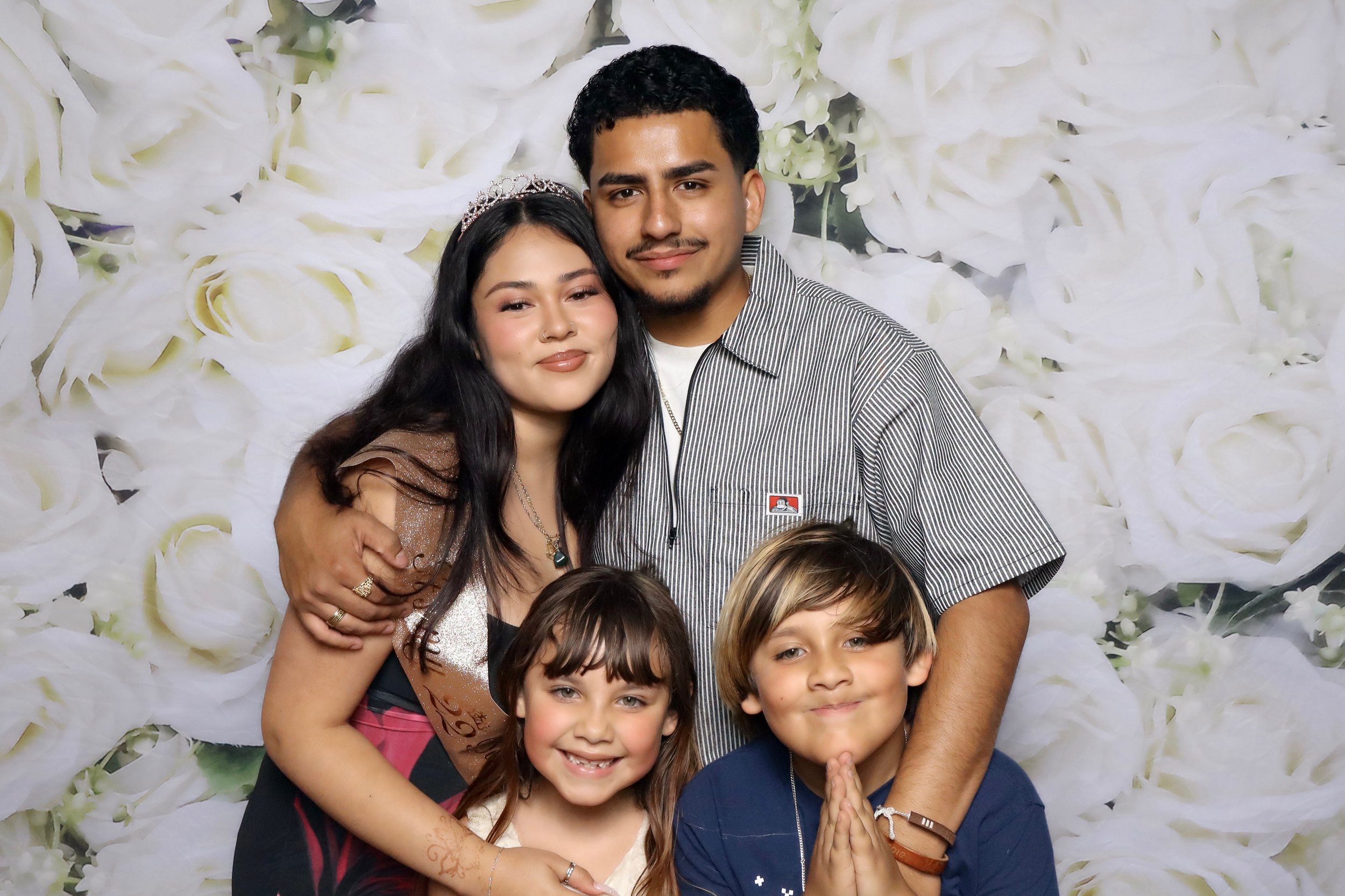 A group of people, including children and adults, posing together in front of a backdrop of white flowers. They are smiling and appear to be happy.