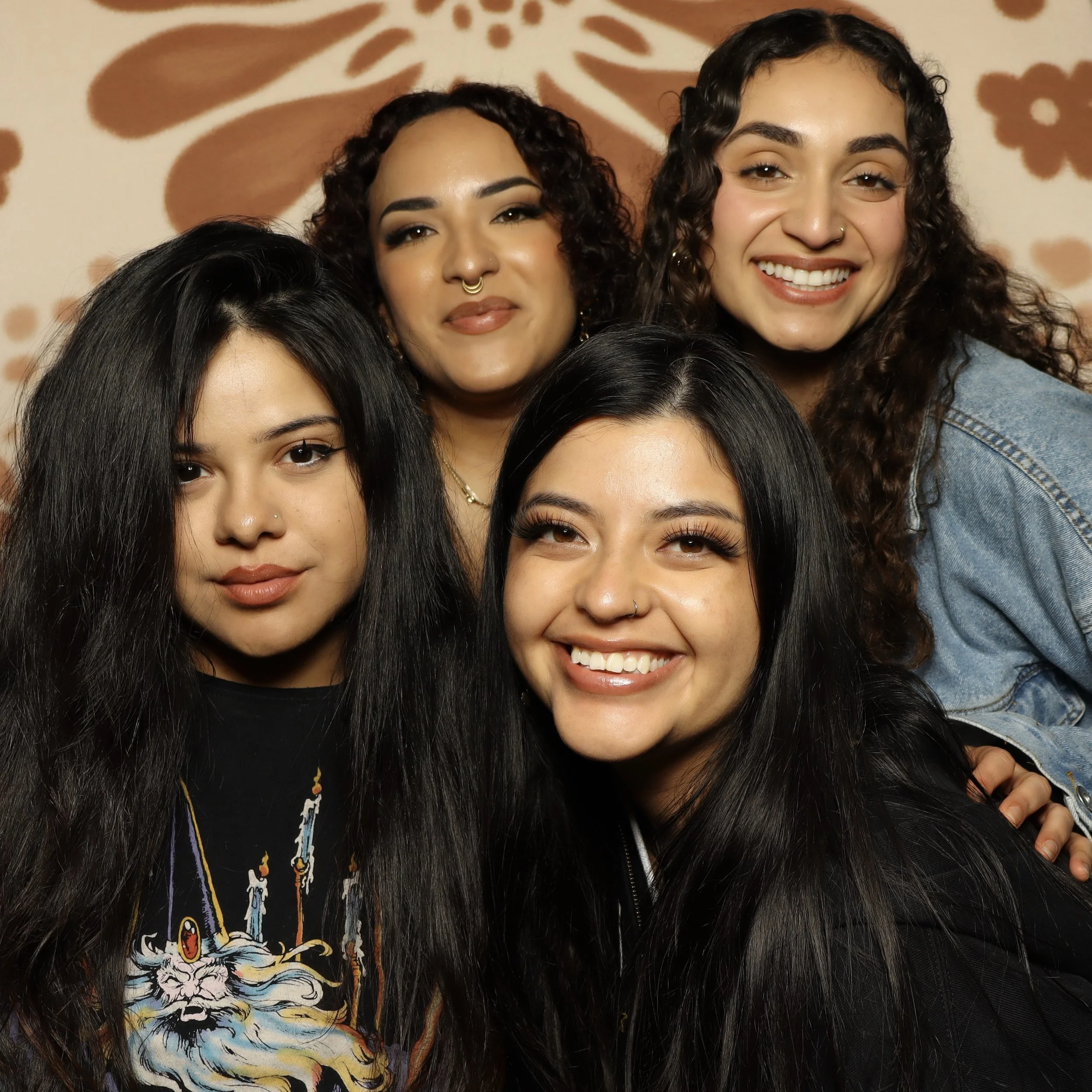 Close-up of multiple women with dark, curly hair and warm smiles, standing close together against a light background with a floral pattern.