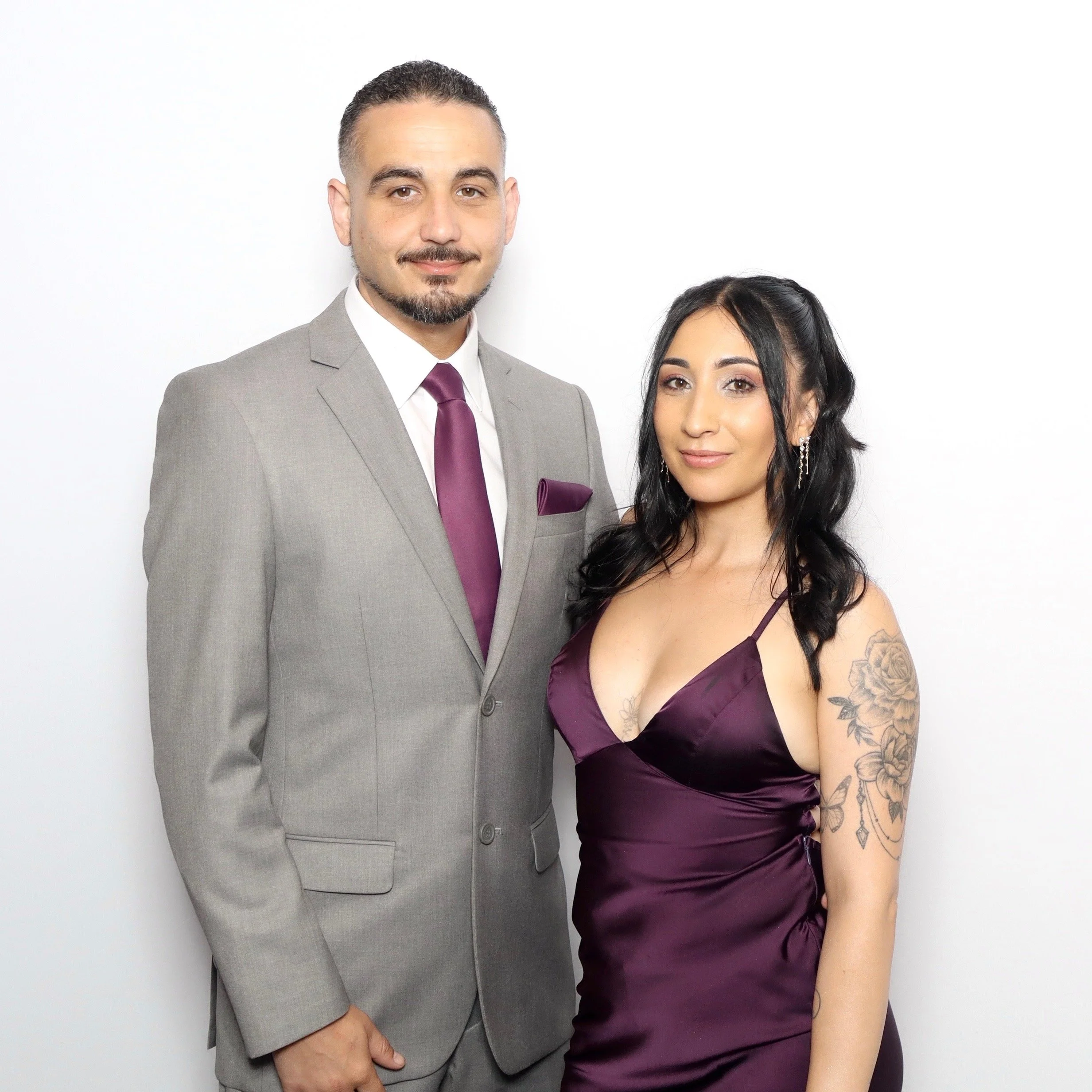 Close-up of a man and woman posing together against a plain white background, dressed formally, with the man in a light gray suit and purple tie, and the woman in a dark purple dress with tattoos on her arm.