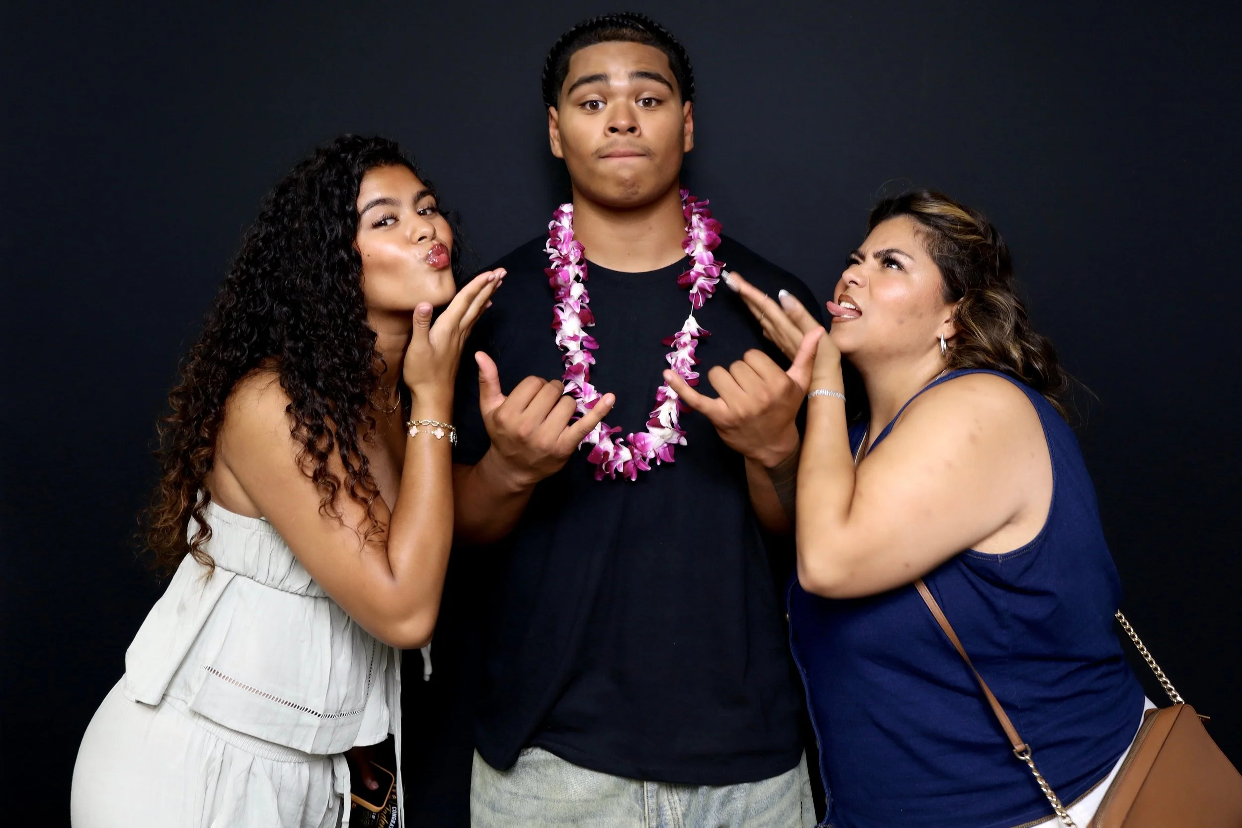 3 young people posed against a black background while the man in the middle wears a lei around his neck.