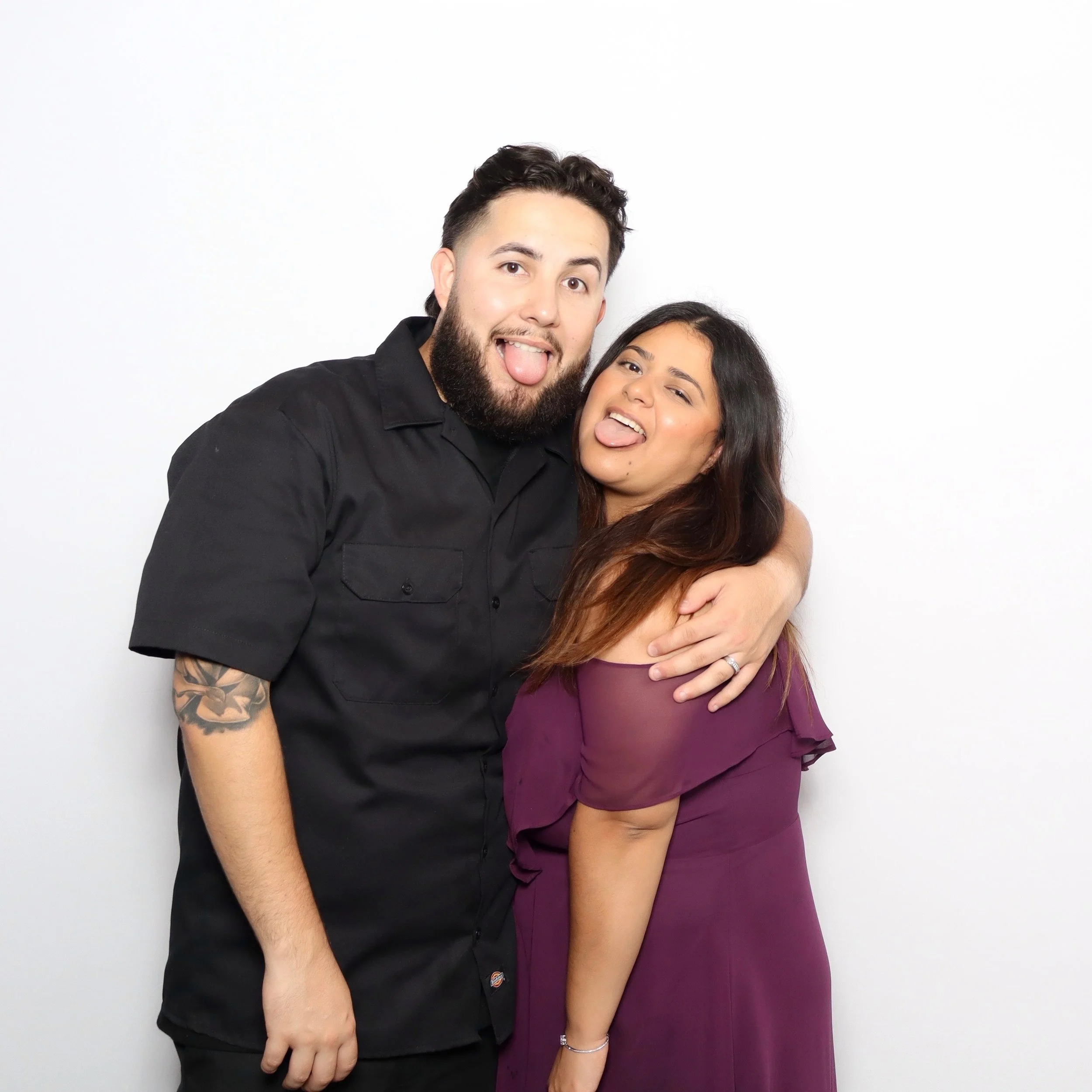 A man and woman making silly faces and sticking their tongues out while hugging against a plain white background.