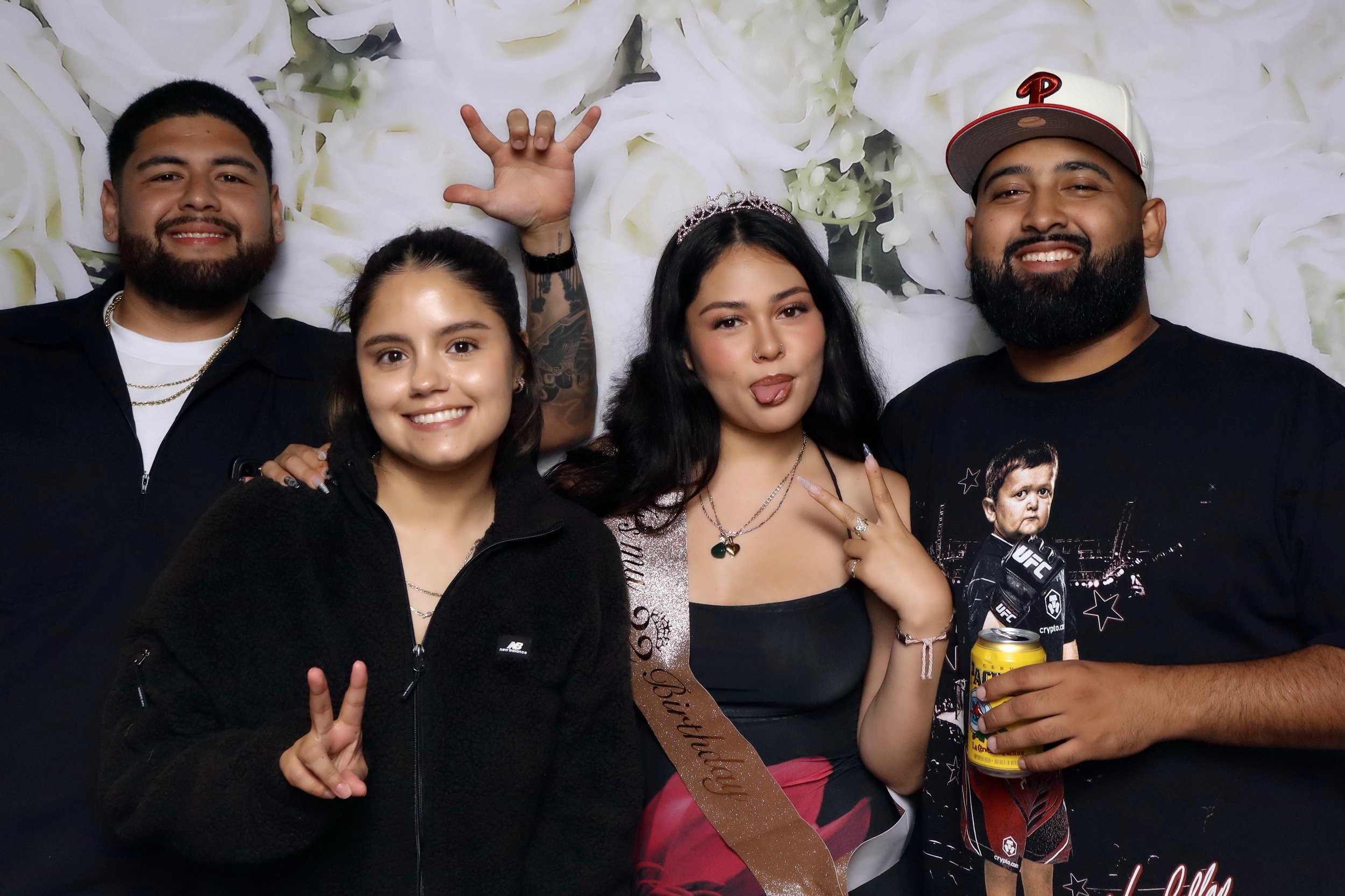 Group of five young adults at a celebration, with three men and two women, some making playful gestures, one woman wearing a tiara and a sash that reads 'Birthday', a man holding a can and a drink, all smiling in front of a floral backdrop.