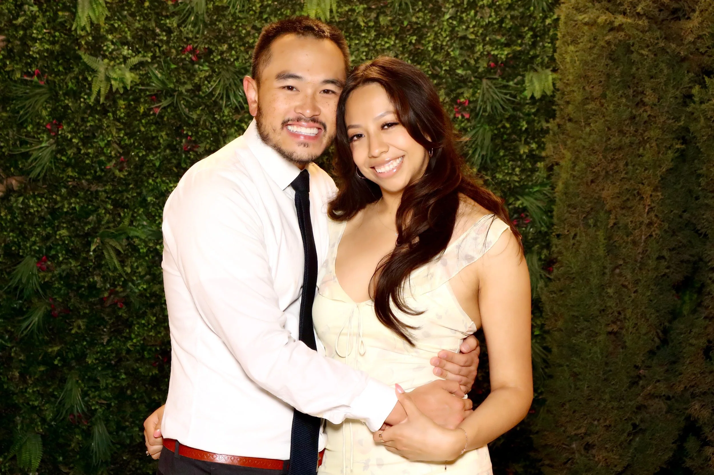A smiling man and woman with light skin, embracing each other in front of a green leafy wall with pink flowers. The man is wearing a white shirt and black tie, and the woman is wearing a light-colored dress.