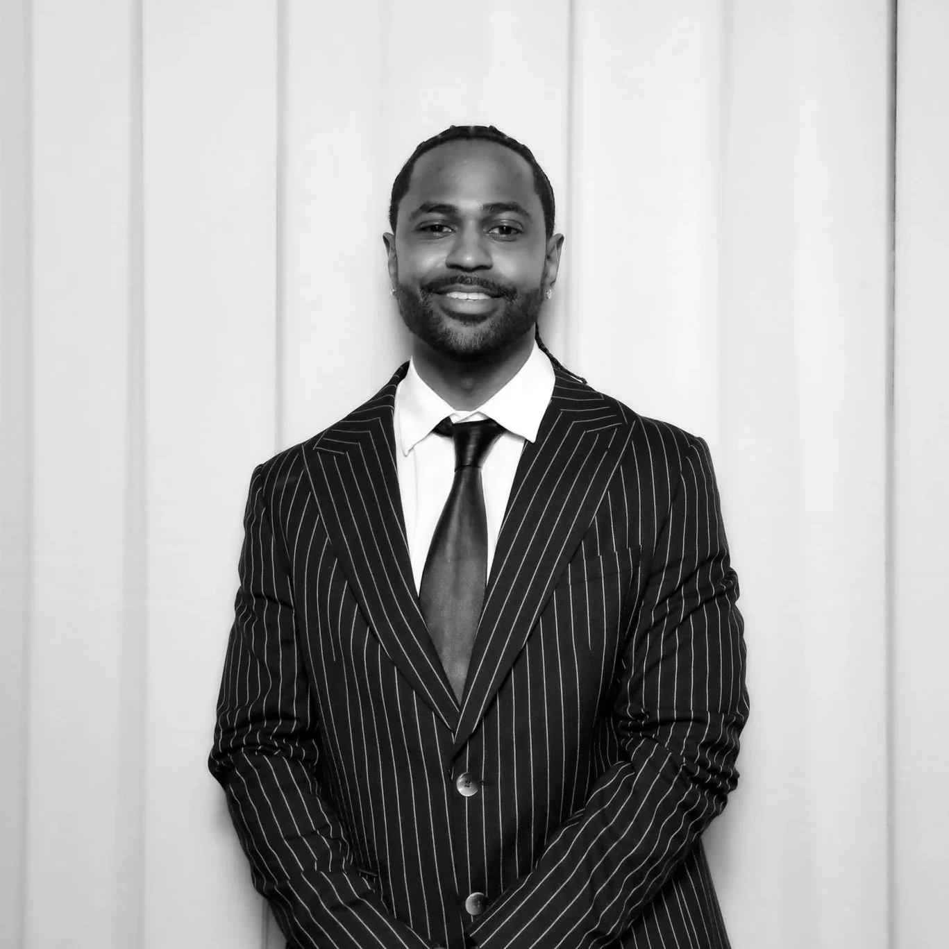 A man in a dark striped suit with a white shirt and black tie, smiling and standing in front of a light-colored wooden wall.