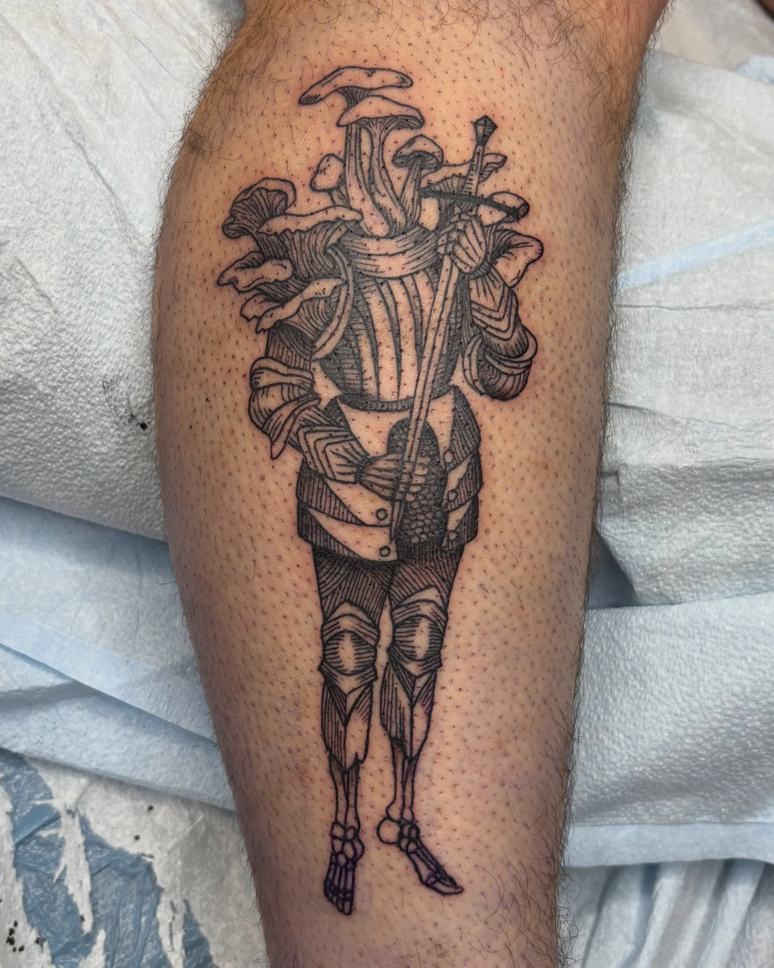 Black outline tattoo of a woman in a dress with mushrooms growing from her head and holding a walking stick, located on a person's leg.