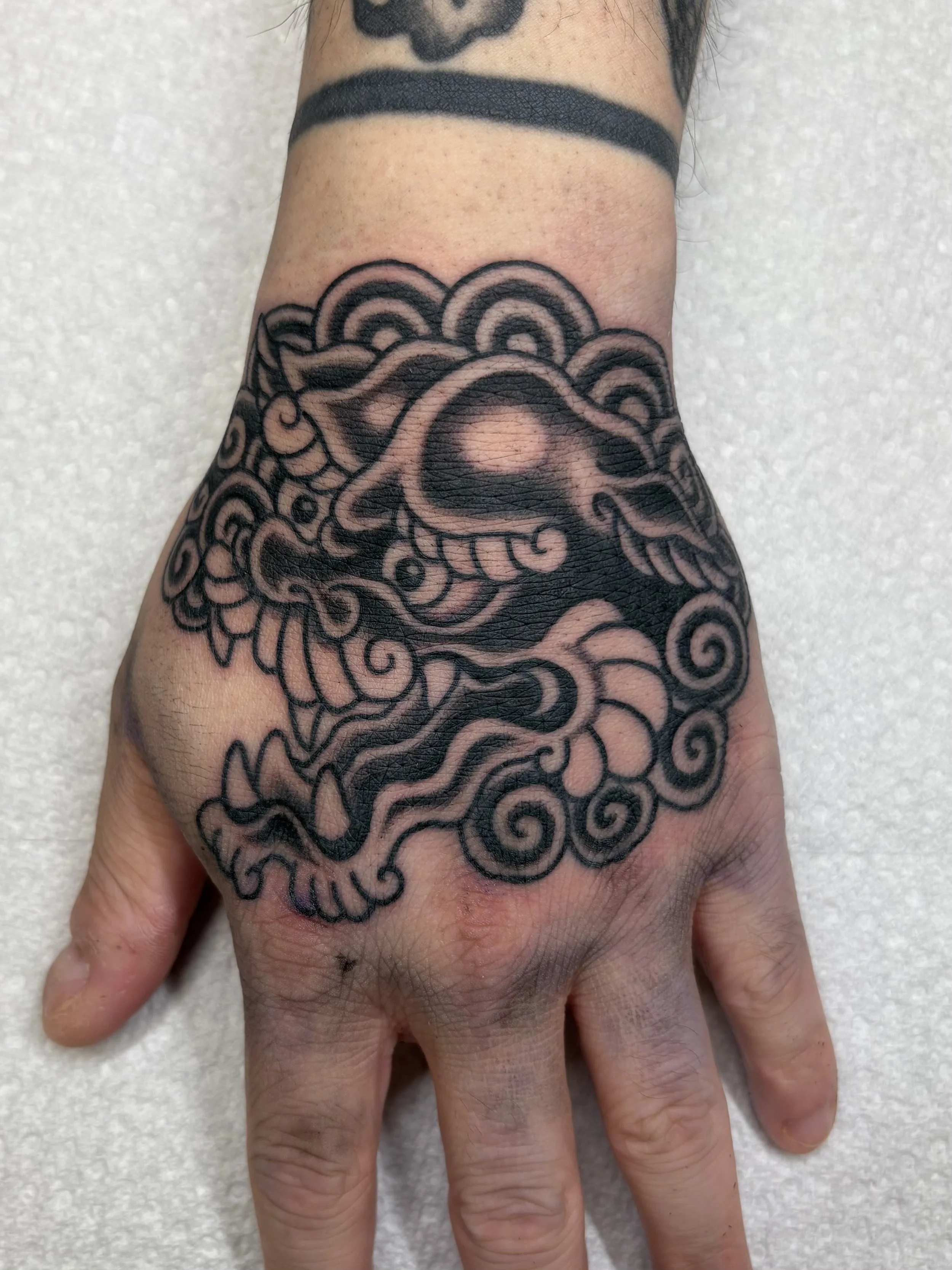 A person's hand with a black ink tattoo of swirling abstract lines and shapes covering the back of the hand and part of the fingers.