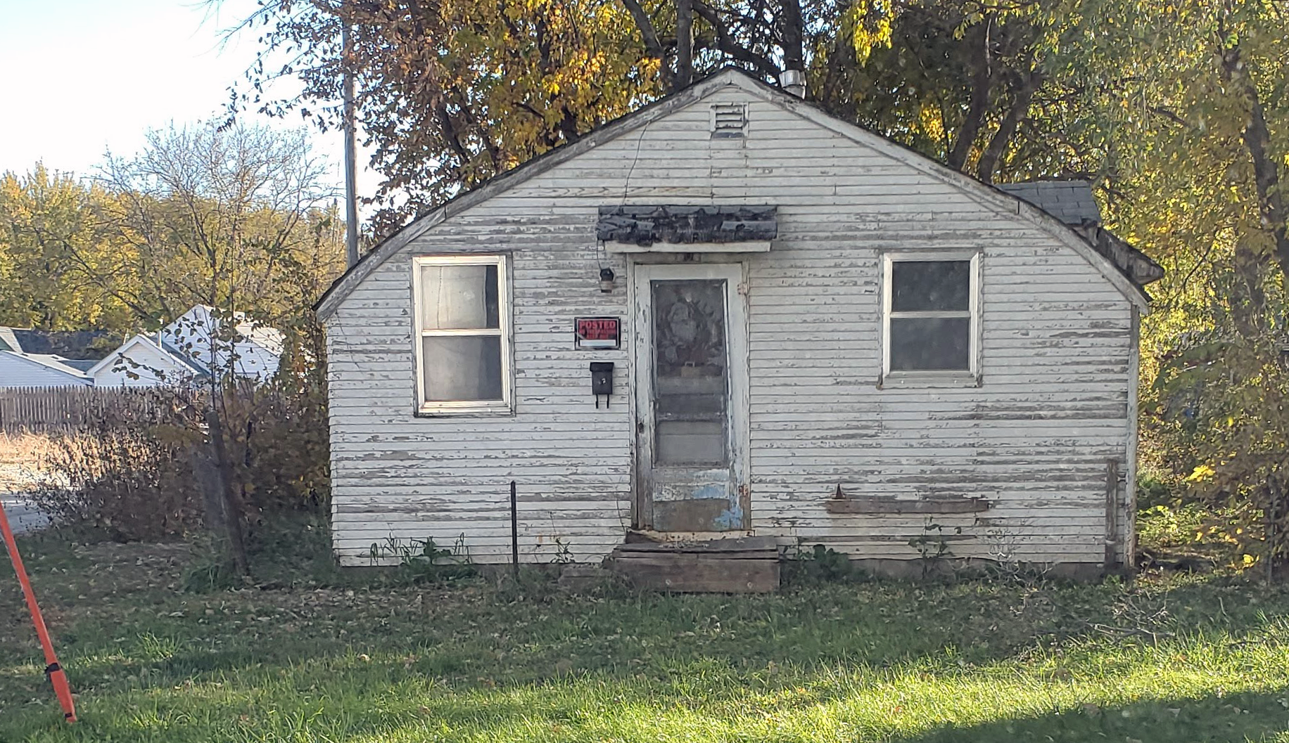 A small, old, weathered house with peeling white paint and a boarded-up front door. Two windows are visible, and there is a "Posted No Trespassing" sign next to the door. The house is surrounded by overgrown grass and trees with fall foliage.