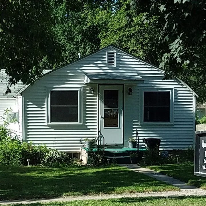 A small white house with two large front windows, a central door, and steps leading up to it. Surrounded by green grass and trees, with a black planter and a sign on the lawn.