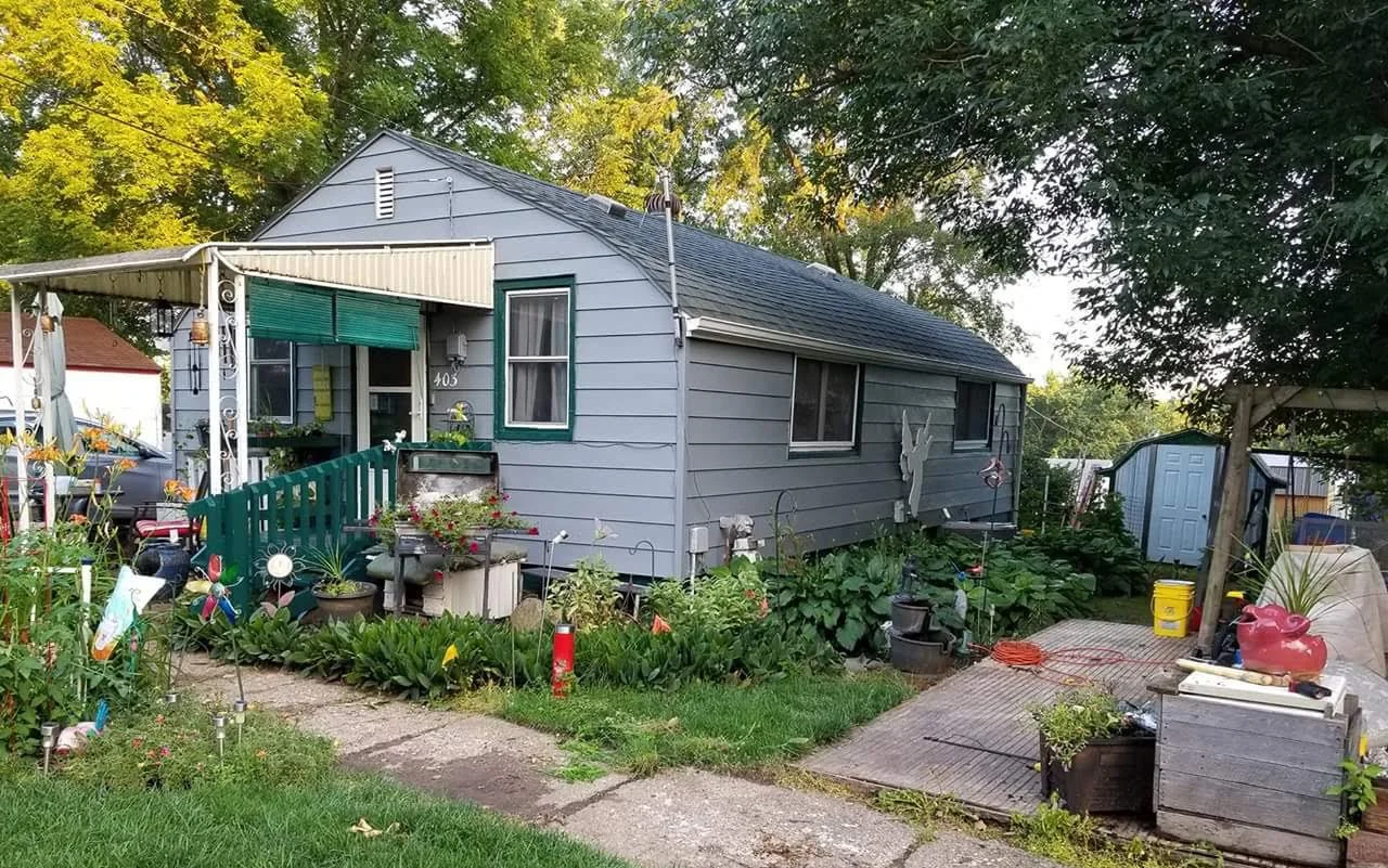 Exterior view of a small, blue house with a green entrance railing, surrounded by a garden with colorful flowers and decorative items. There is a small shed to the right and trees in the background.
