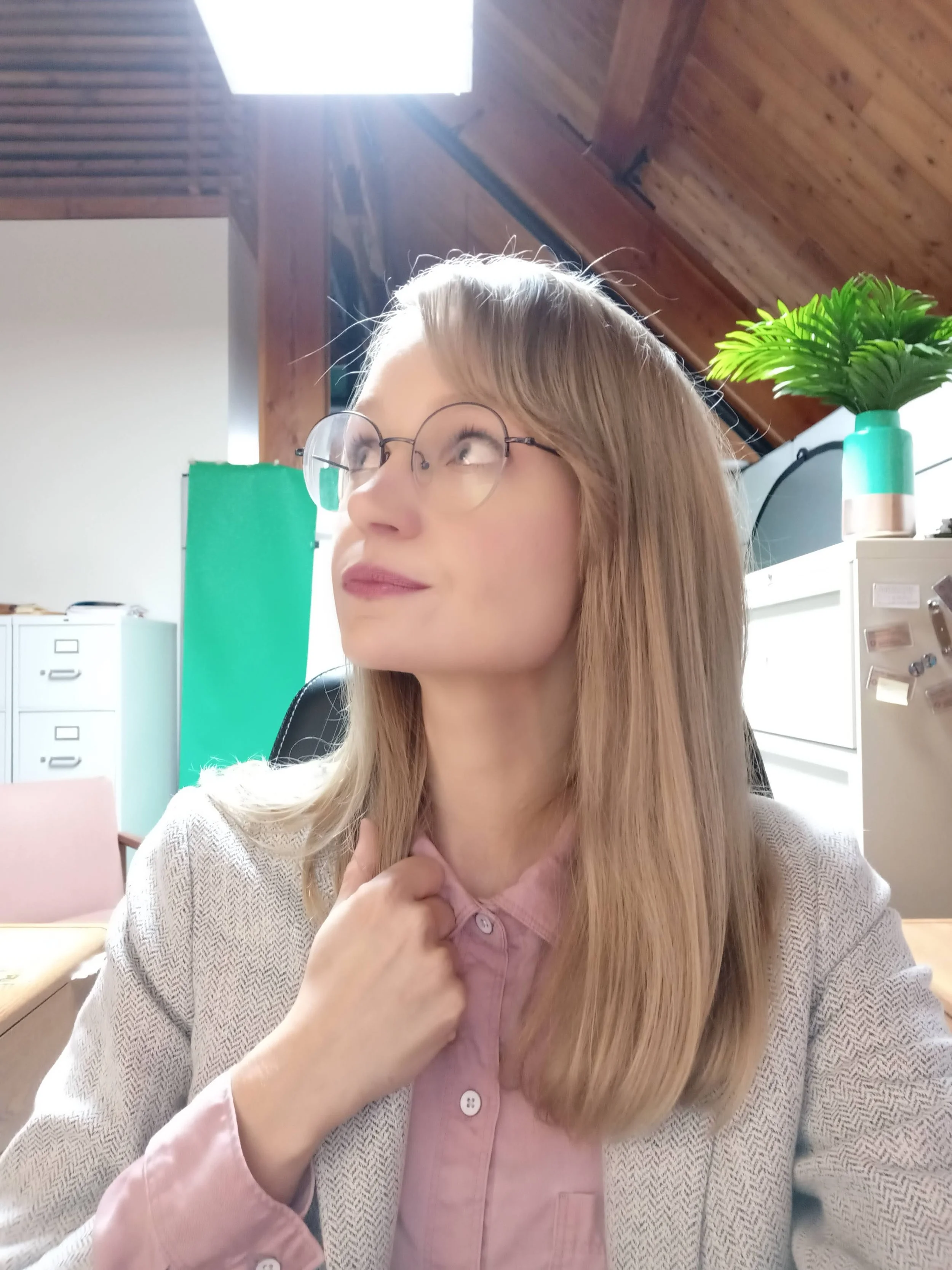A woman with long blonde hair, wearing glasses and a beige jacket over a pink shirt, sitting in a room with wooden ceilings and modern furniture. She has a thoughtful expression and is touching her collar.