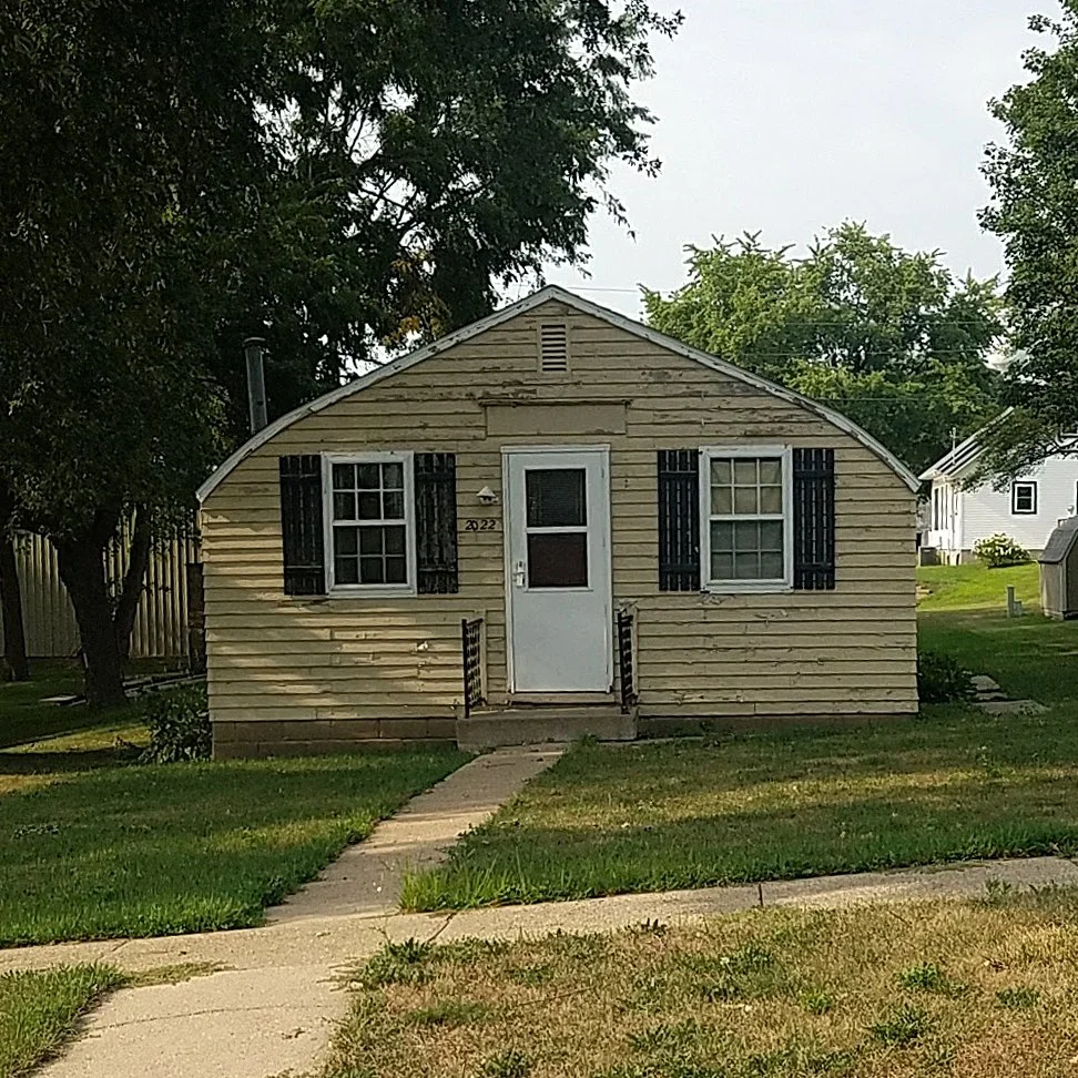 A small, weathered house with peeling beige siding, black shutters, a white door, and a small porch with steps. The house is flanked by a lawn and trees.