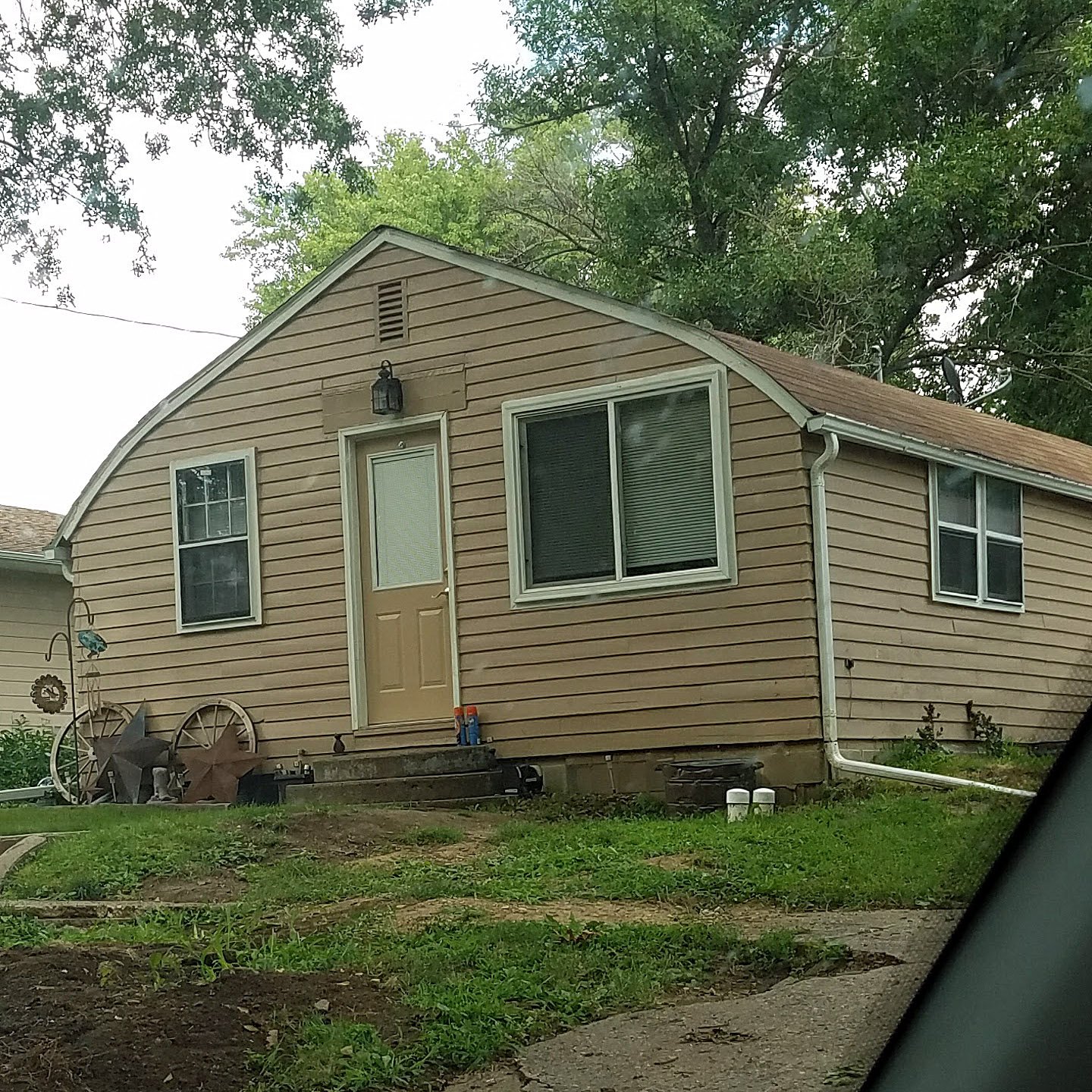 A beige house with white trim, a small front porch, and a brown shingled roof. Two front windows, a door with a small window, and decorative outdoor items in the yard. Green trees in the background.