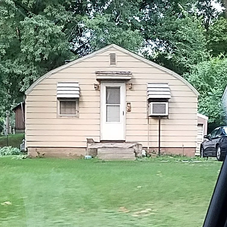 Small beige house with a curved roof, two small windows with white shutters, an air conditioning unit, and a white door with steps, surrounded by green trees and grass.