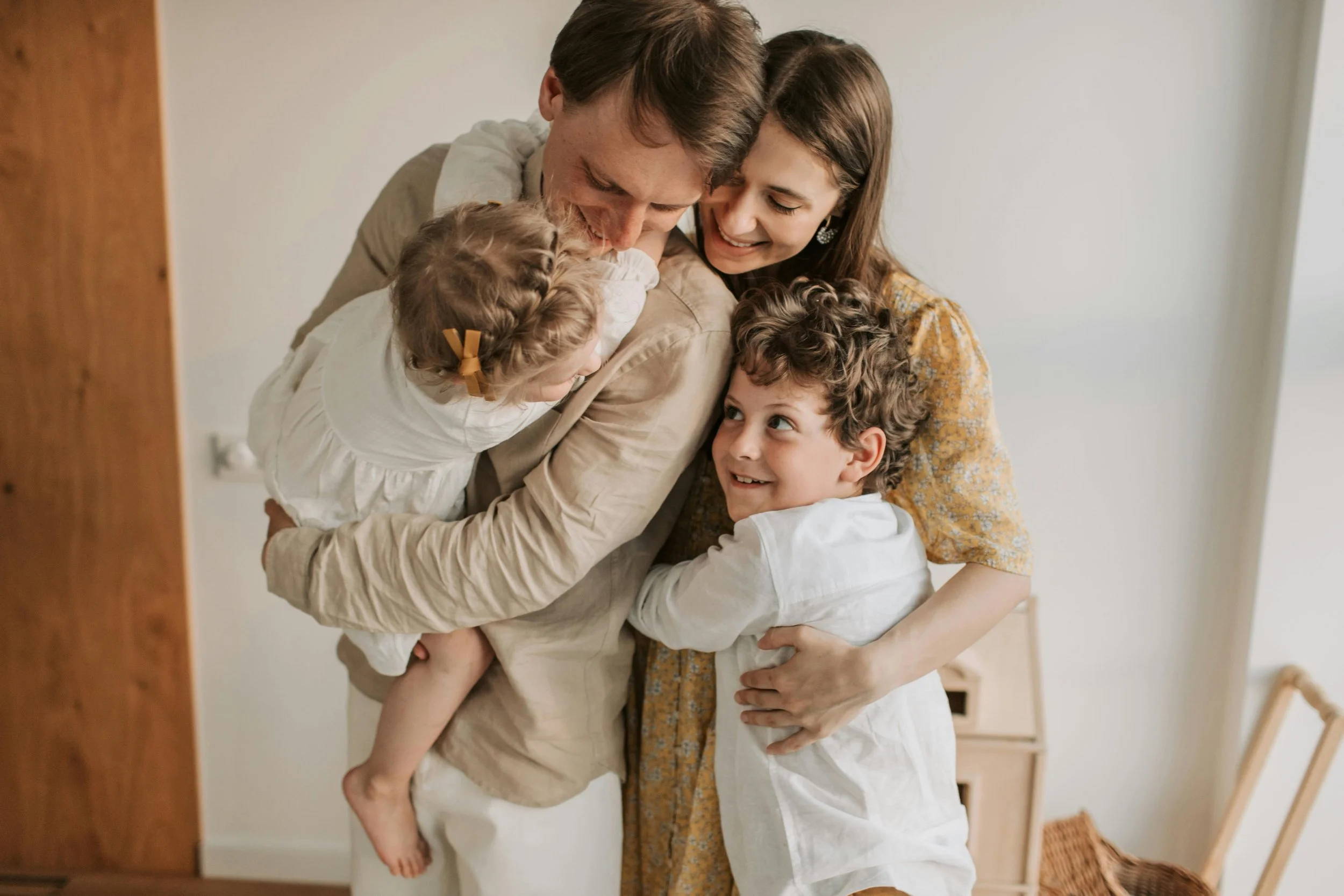 A family of four hugging each other indoors, smiling and displaying affection.