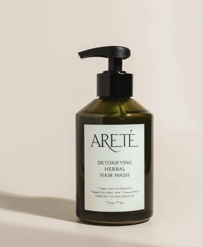 A dark green bottle of ARETÉ Detoxifying Herbal Hair Wash with a black pump dispenser, placed on a light-colored surface with a plain background.