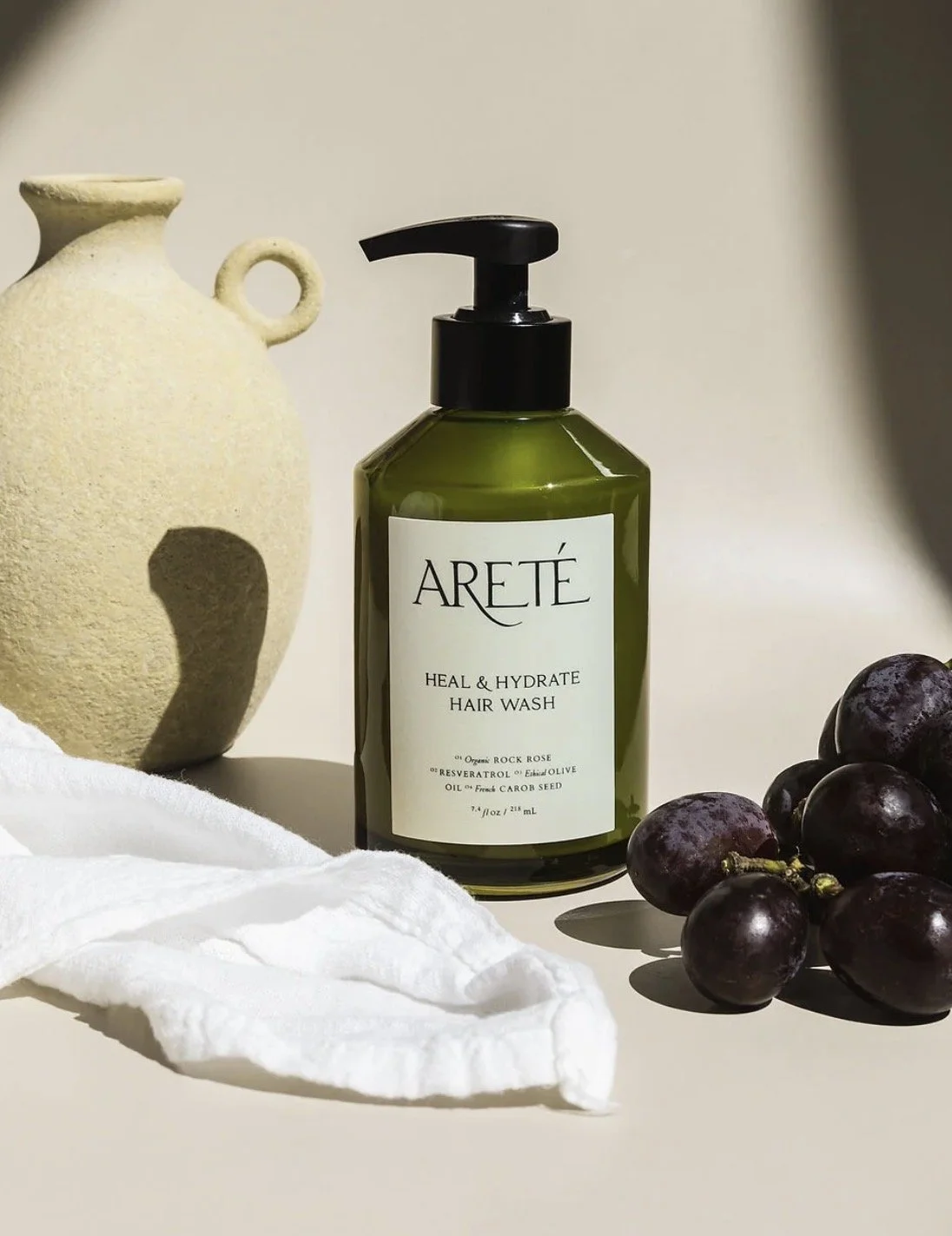 A green bottle of ARETÉ Heal & Hydrate Hair Wash with a black pump, a bunch of grapes, a white cloth, and a beige ceramic vase on a light surface with a plain background.