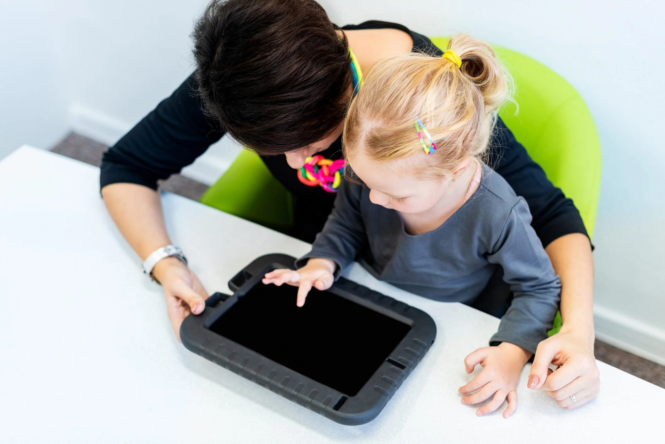 A woman and a young girl are sitting at a white table, looking at an AAC device, communicating together.