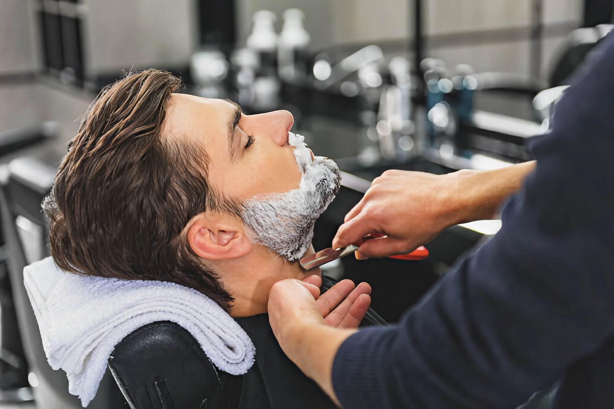A man with short brown hair is lying back at a barber shop chair with a towel around his neck, receiving a beard trim from a barber. The barber is holding clippers and is trimming the man's beard while the man's eyes are closed.