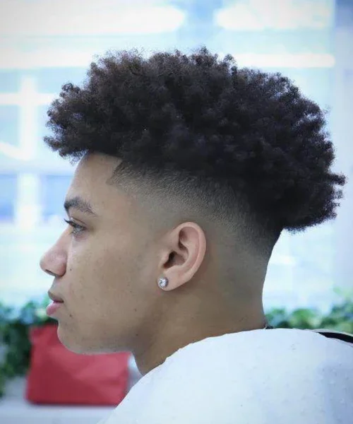 Side profile of young man with curly afro hairstyle, shaved fade hairstyle, wearing stud earring, indoors with a blurred background.
