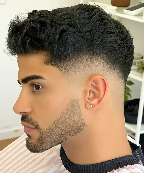 Side profile of a young man with a textured curly hairstyle, faded haircut, and a groomed beard, wearing a small earring.